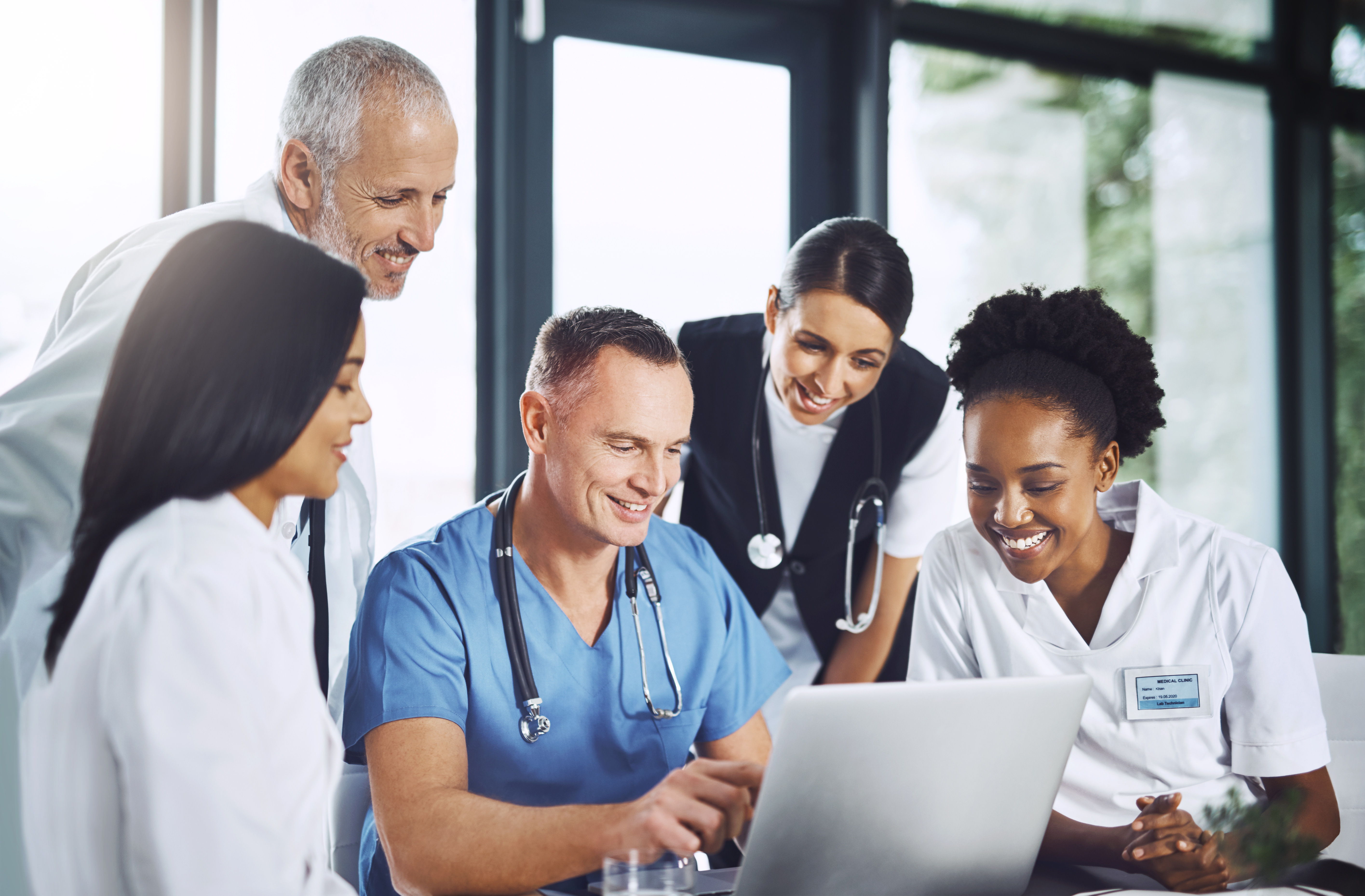 Group of clinicians in scrubs and white coats huddled together looking at a laptop