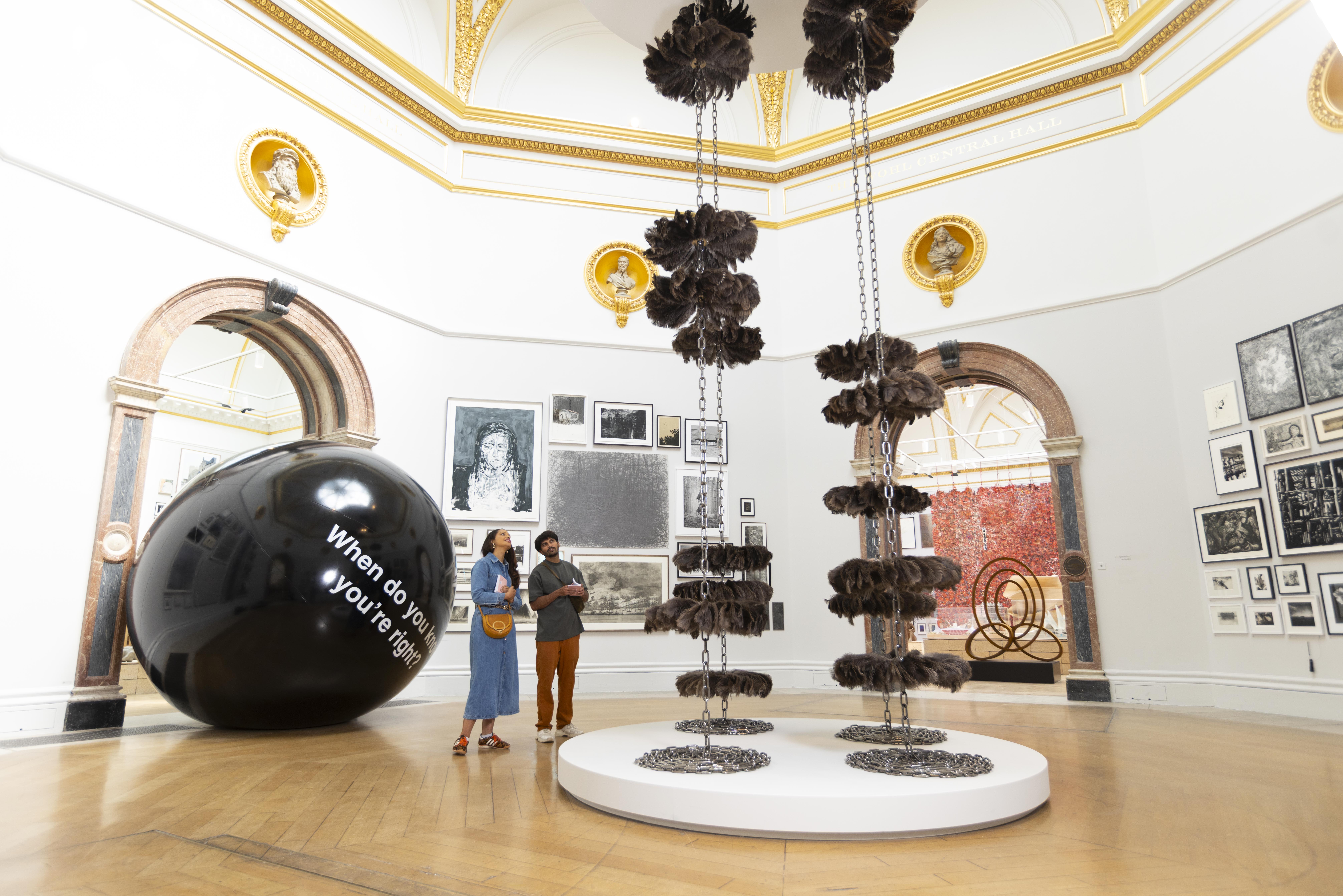 Two people stand in a gallery, looking at two large installations. The first is a shiny black sphere with the text 