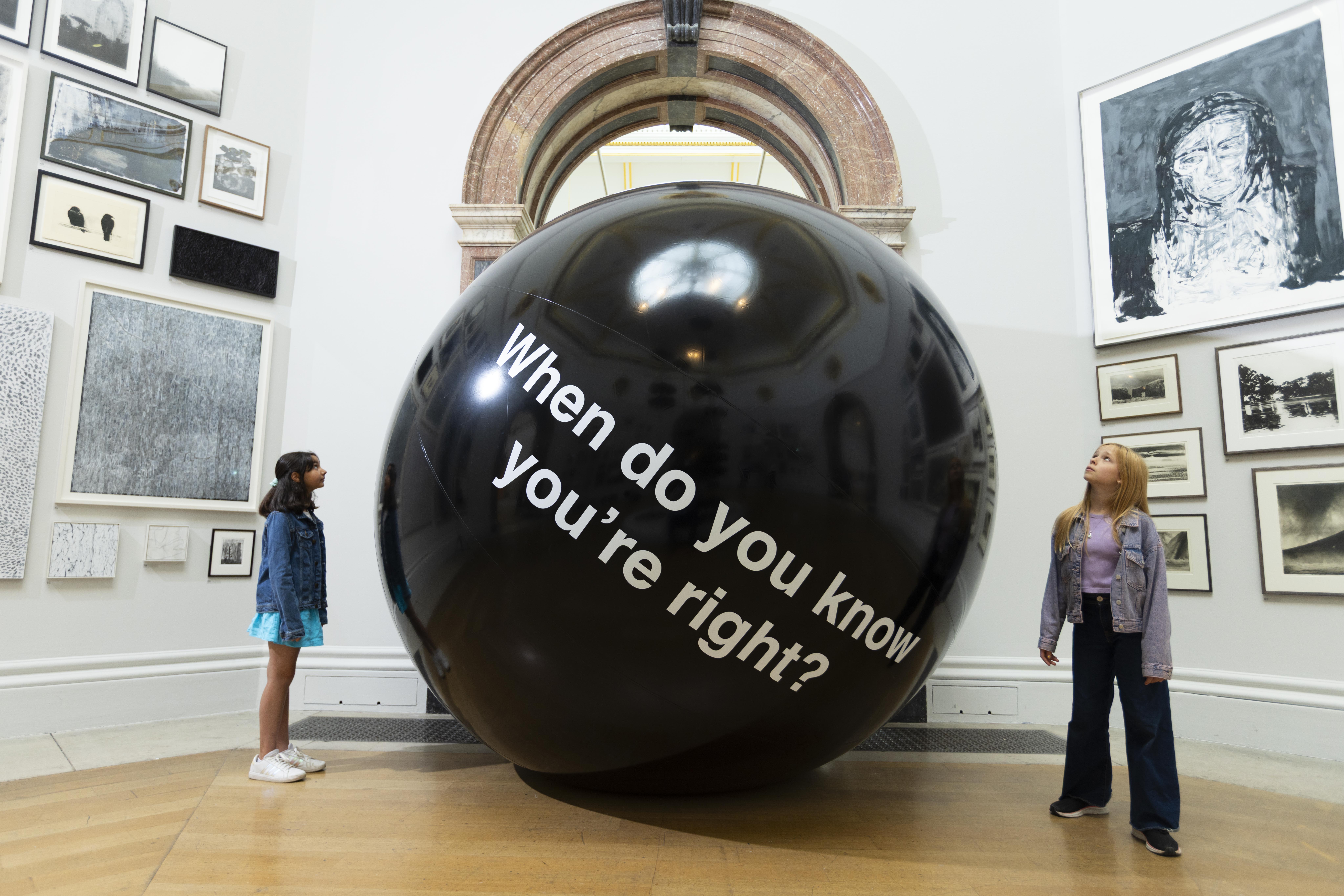 A child looks up at a massive, glossy black sphere. The sphere has white text on it that reads, 