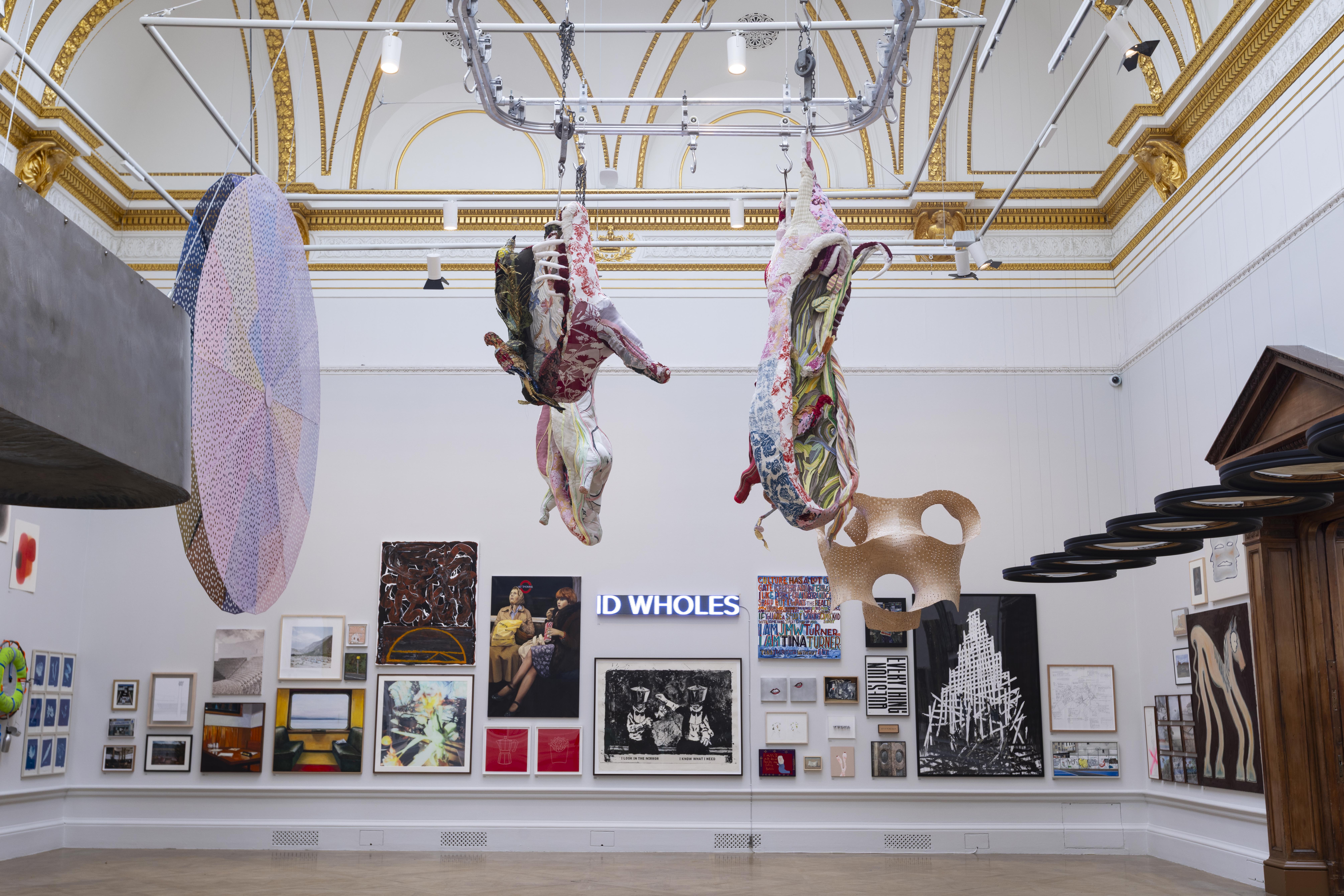 
An installation by Tamara Kostianovsky hangs from the ceiling of a gallery. The two main pieces resemble animal carcasses, but they're made from multicolored textile scraps. They hang from a metal track system like those found in a butcher's shop. In the background, there's a gallery wall filled with various other artworks.
