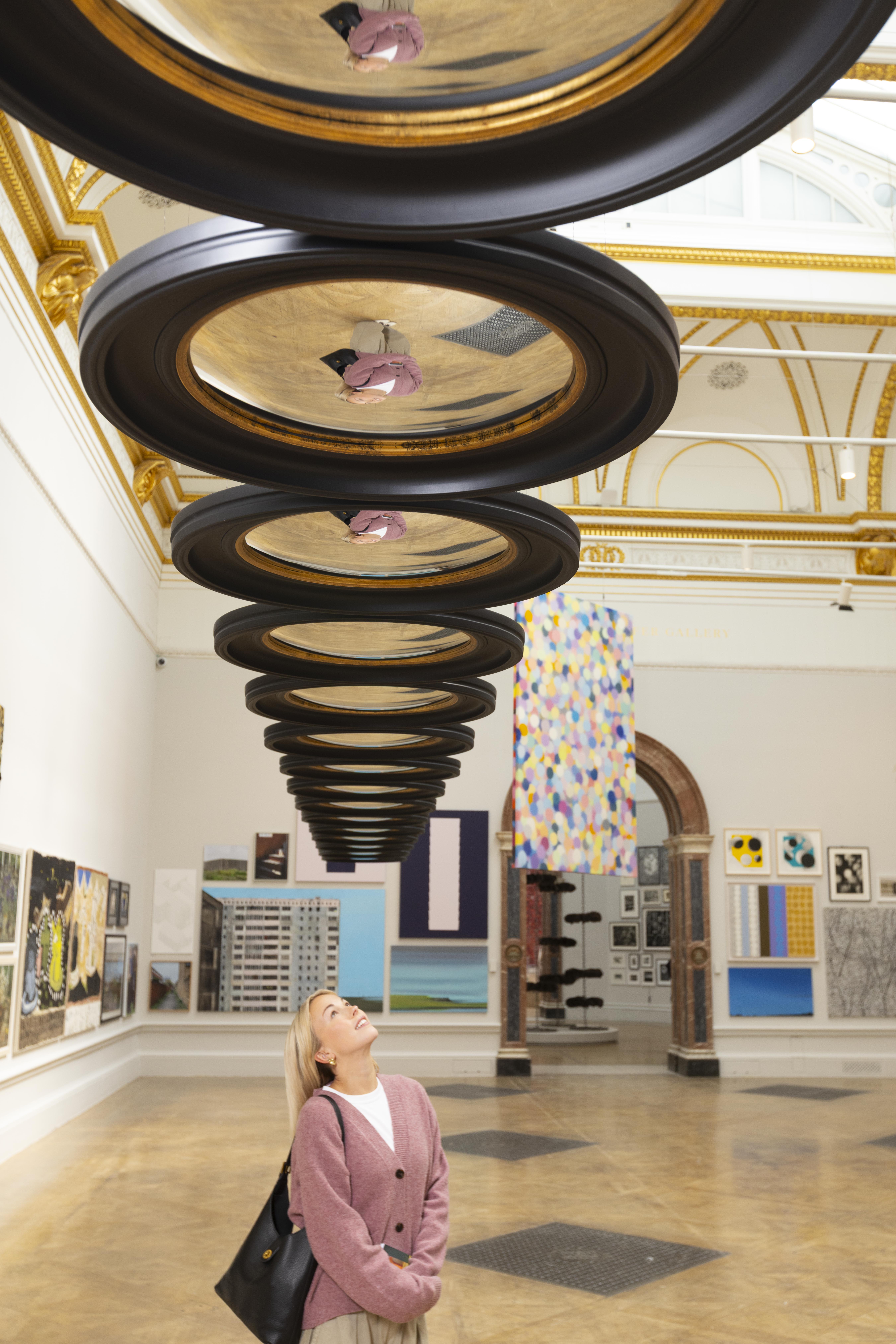 A blonde woman looks up at a long row of what appear to be stacked, reflective picture frames suspended from the ceiling. The frames are part of an art installation by Cornelia Parker at the Royal Academy of Arts Summer Exhibition. Other artworks, including a large, colorful hanging piece and framed prints, are visible on the gallery walls in the background.