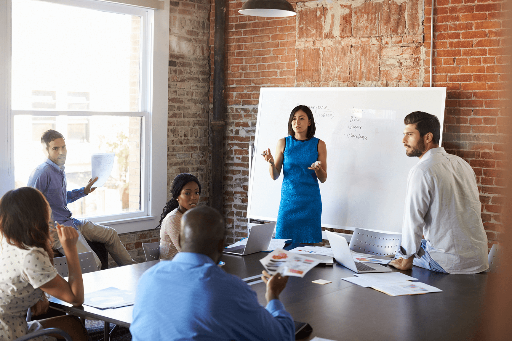 A businesswoman in front of a dry erase board pitching something to a group of workers around a table