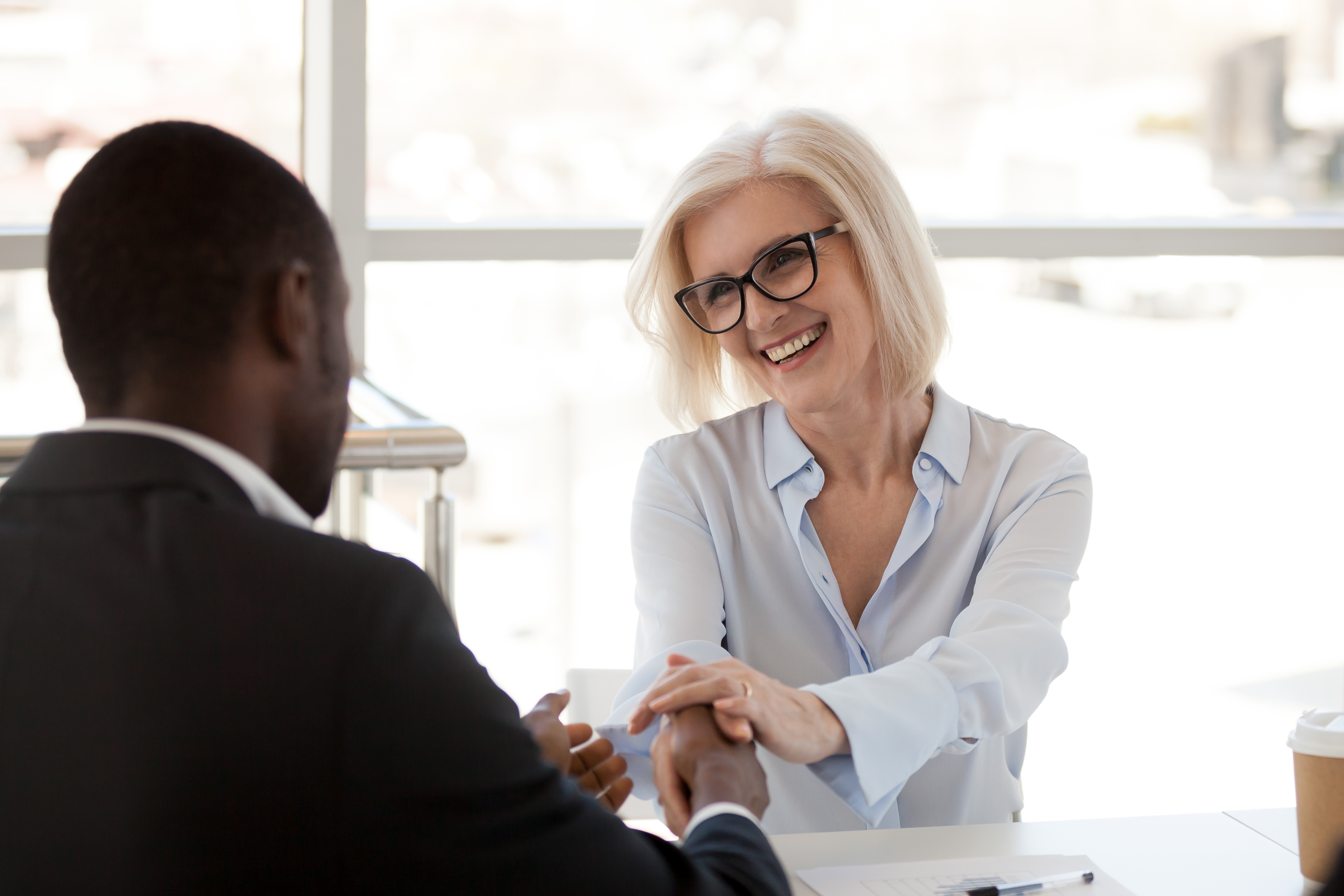 Smiling happy middle aged female caucasian businesswoman holding shaking hand of african american male partner thanking for help at meeting, business loyalty, trust, respect and gratitude concept