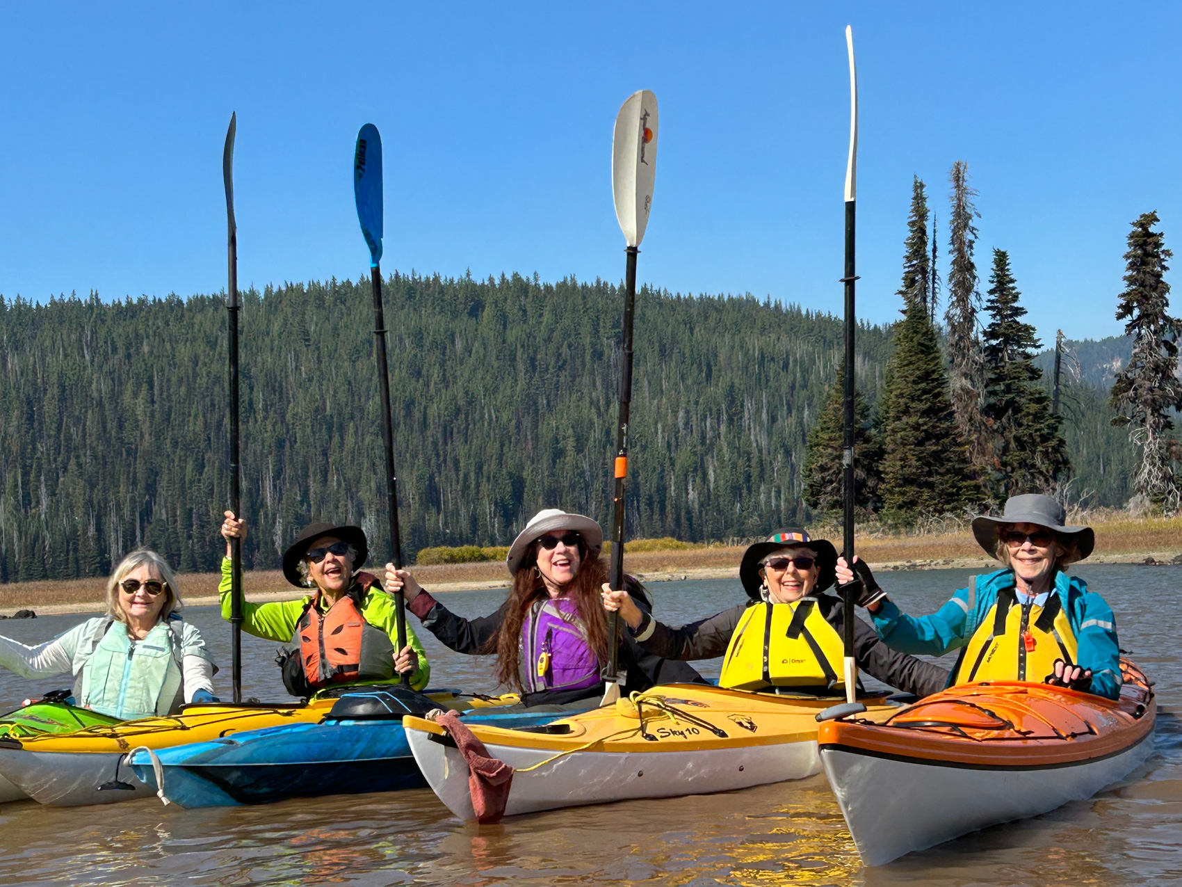 Six women in their 60s are sitting in colorful kayaks, holding their paddles in the air and smiling.