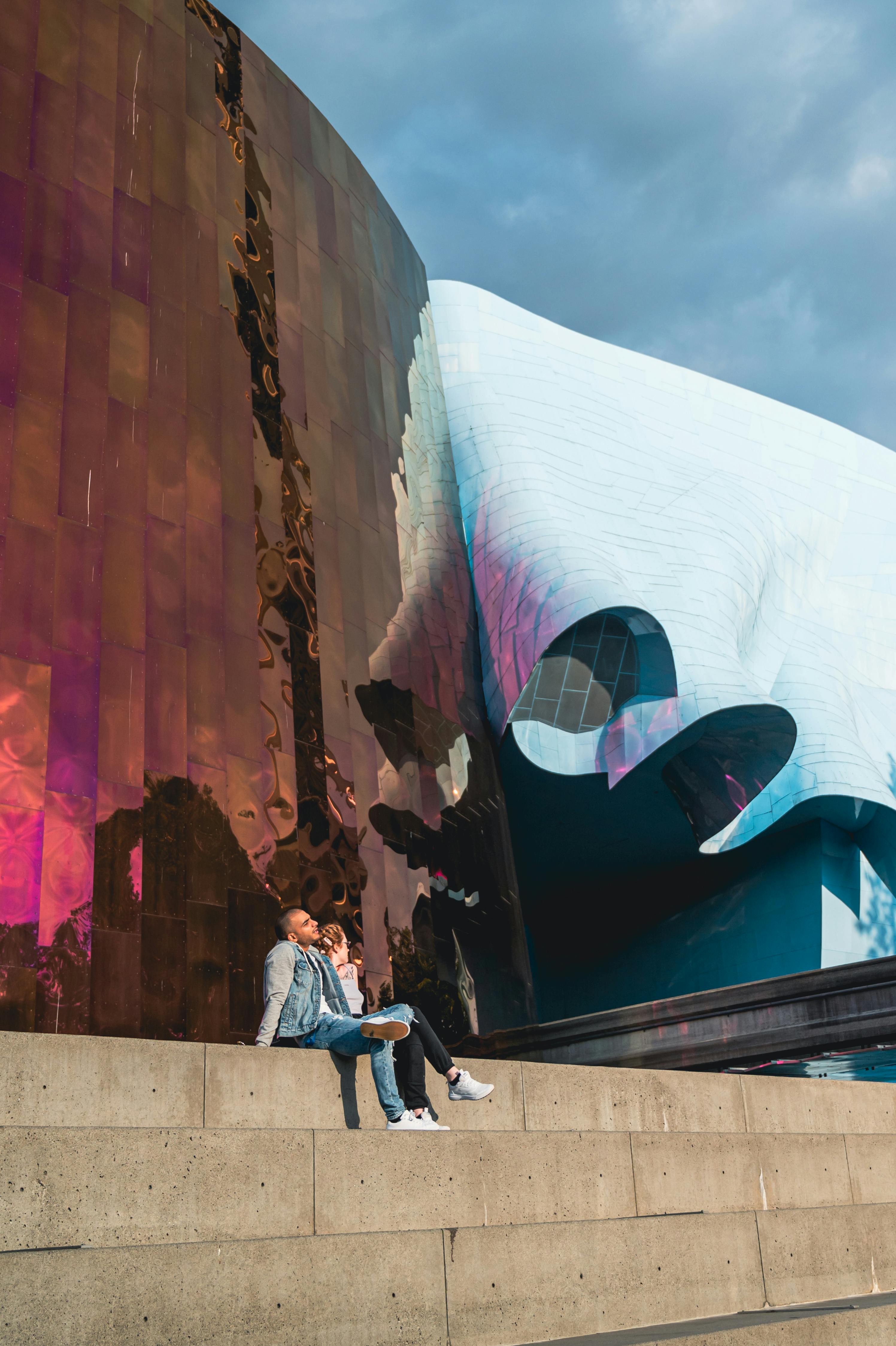 Two people sitting on the outdoor steps of the Museum of Pop Culture, a curvy building clad with colorful metal sheets.