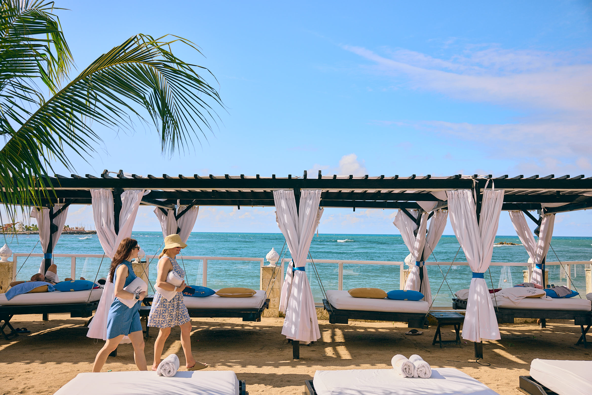 Two women holding white towels, walking along a row of cabanas at the beach