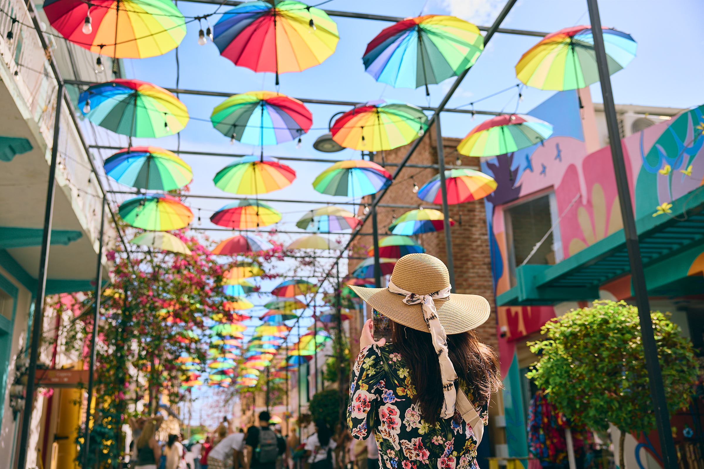 Woman taking a photo of an alley with dozens of rainbow umbrellas suspended in the air above her