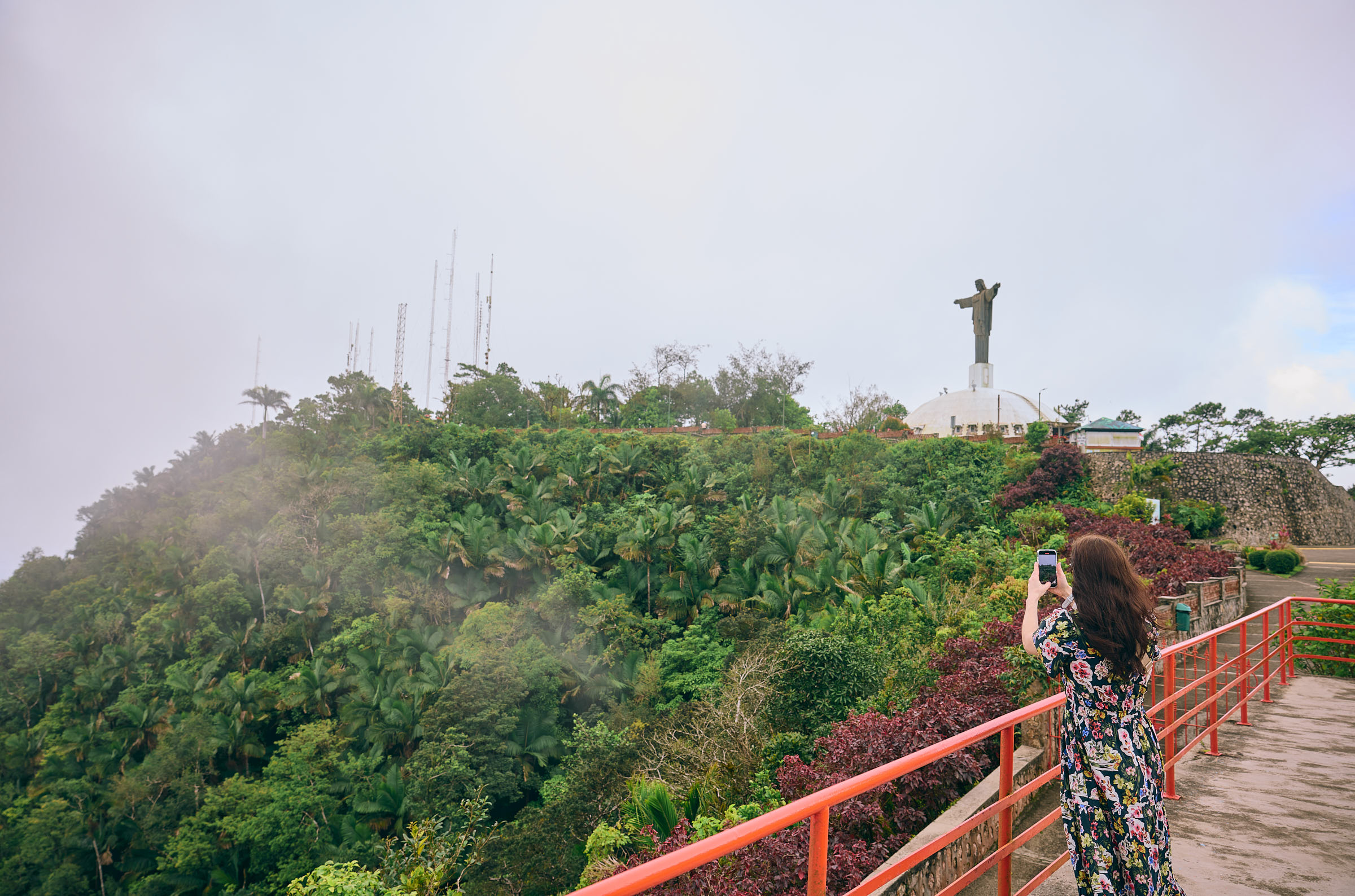 Woman at the top of a hill, standing near a railing and taking a photo of a large statue of Jesus Christ with his arms outstretched