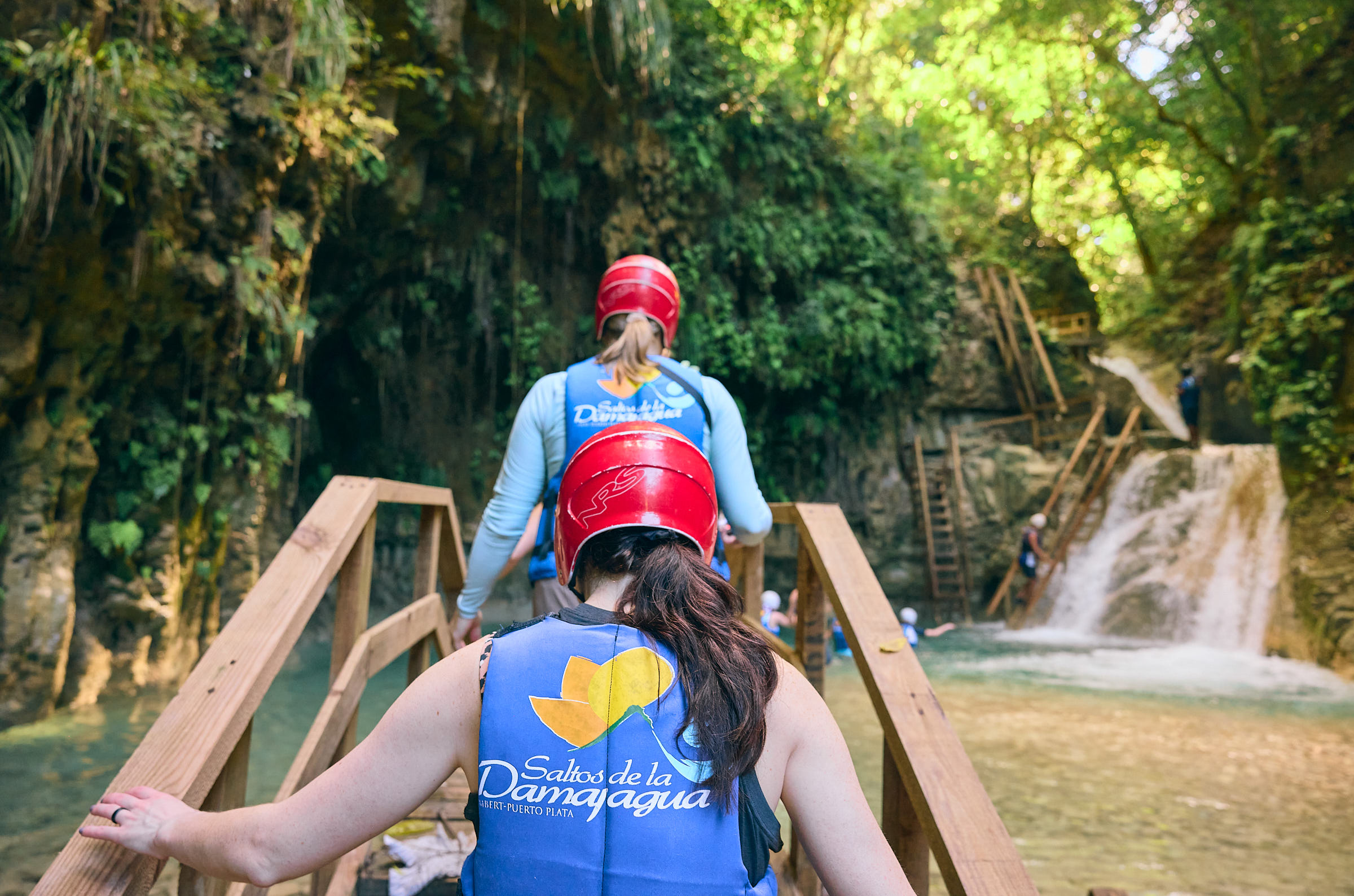 Two women in swimsuits, floatation vests, and helmets climbing up a set of wooden stairs in a jungle