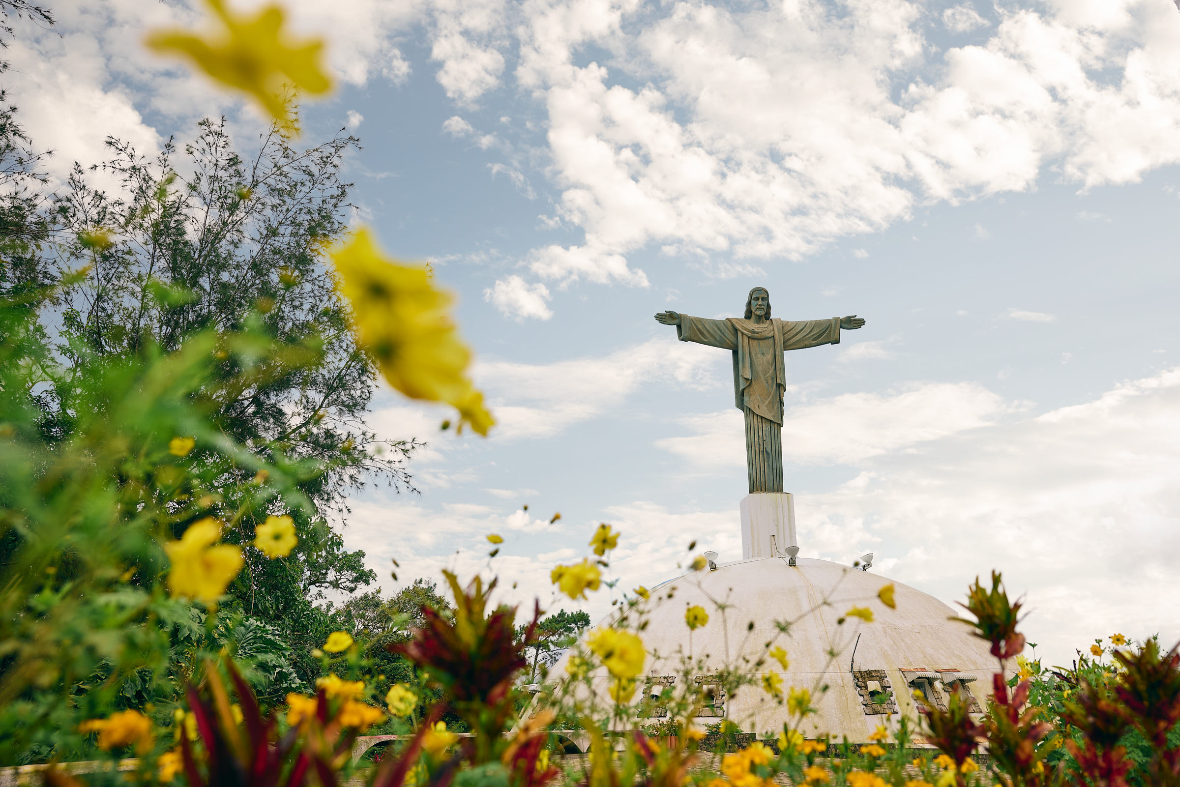 Large statue of Jesus Christ with his arms outstretched at the top of a hill, with flowers framing the shot 