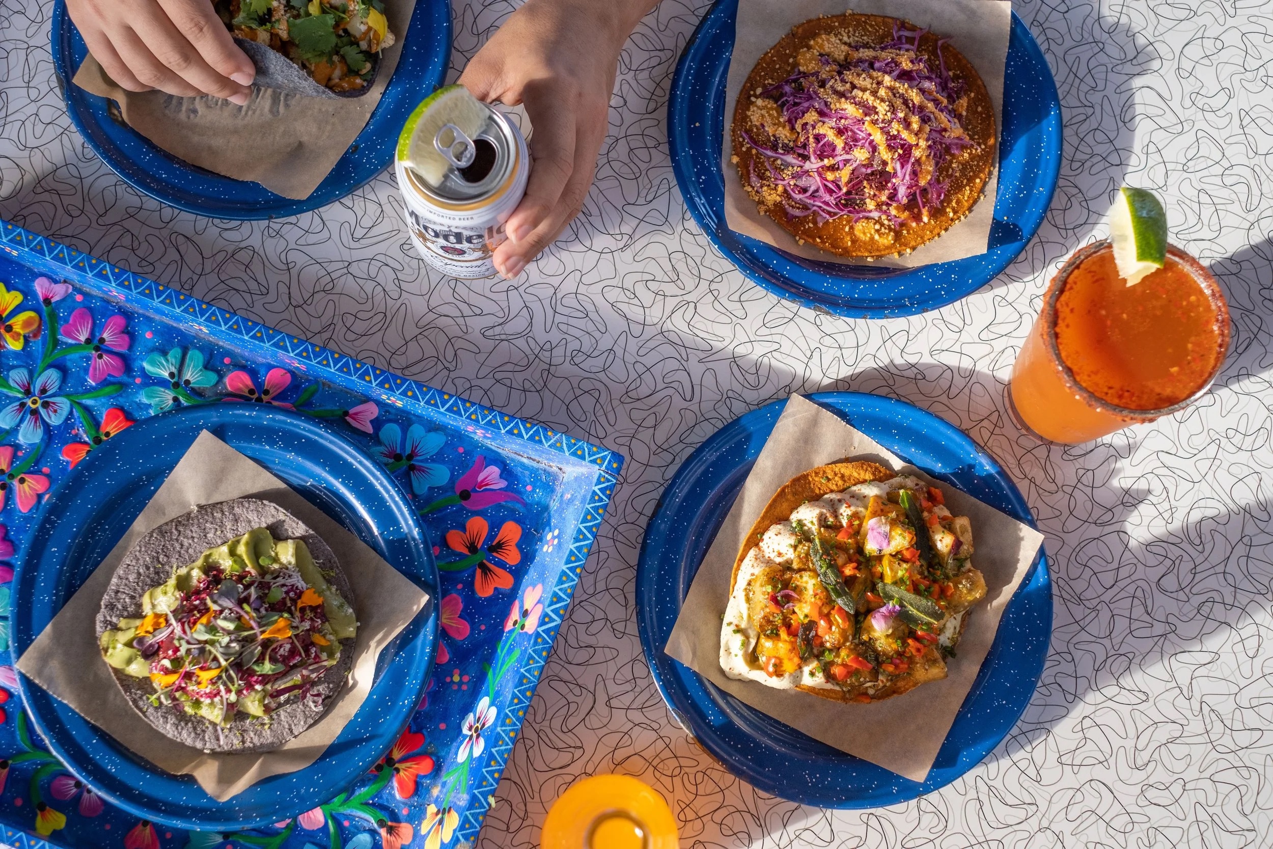 Overhead view of hispanic food on blue plates, along with someone holding a Modelo on a formica table