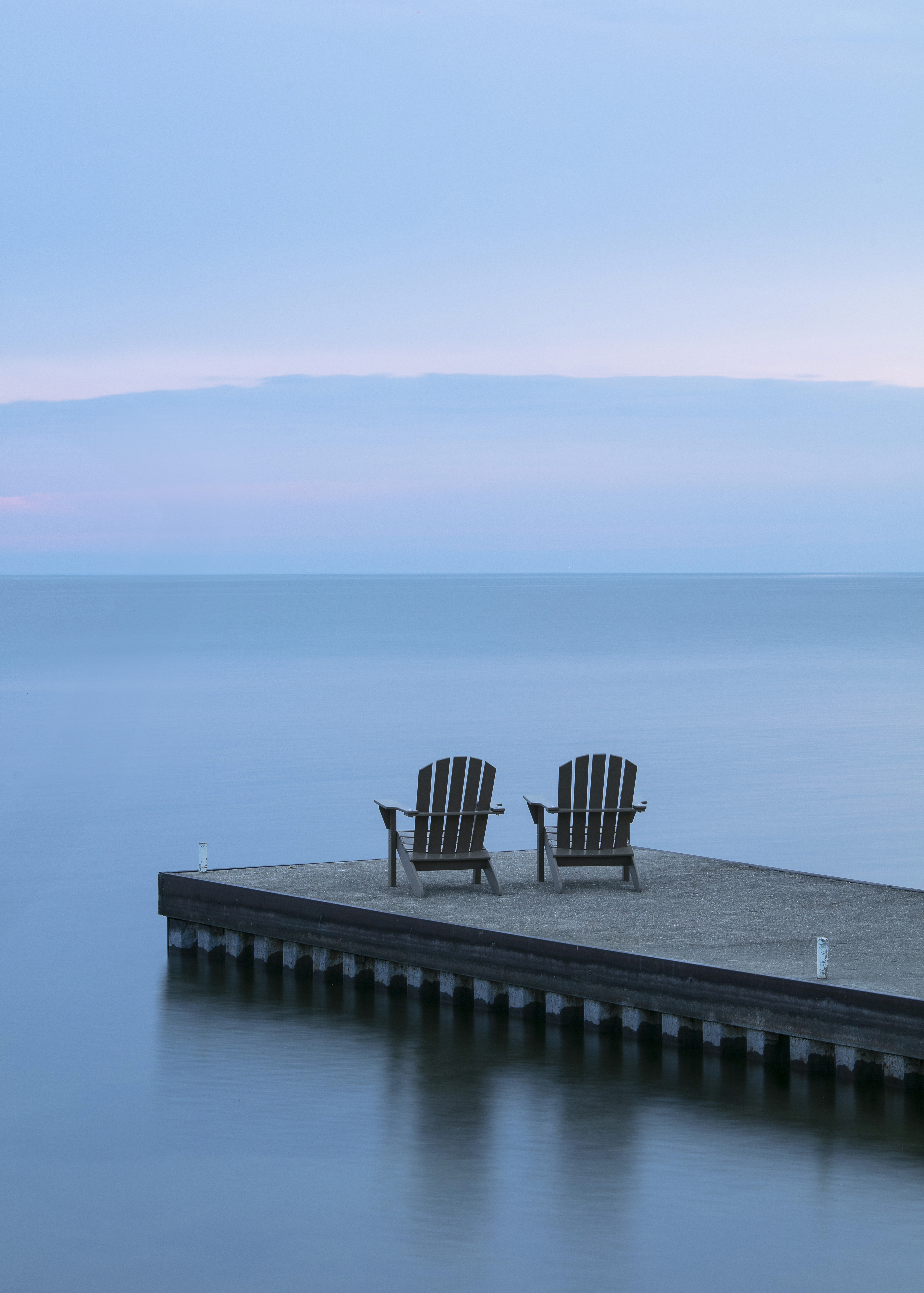 Two Adirondack chairs at the end of a lake dock in dusky purple light