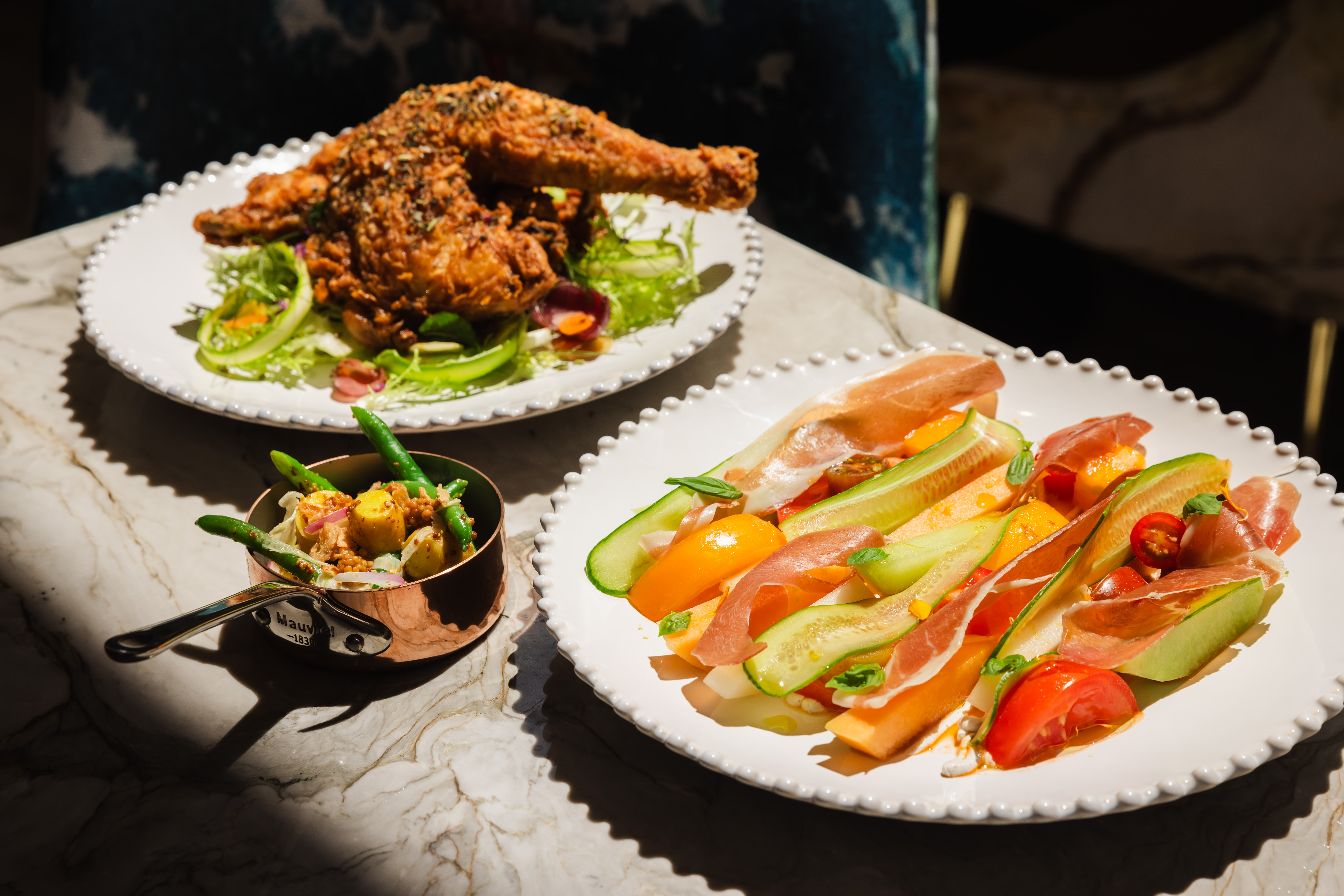 Elegant white plates with fried chicken, sliced cucumber, and prosciutto on a marble countertop