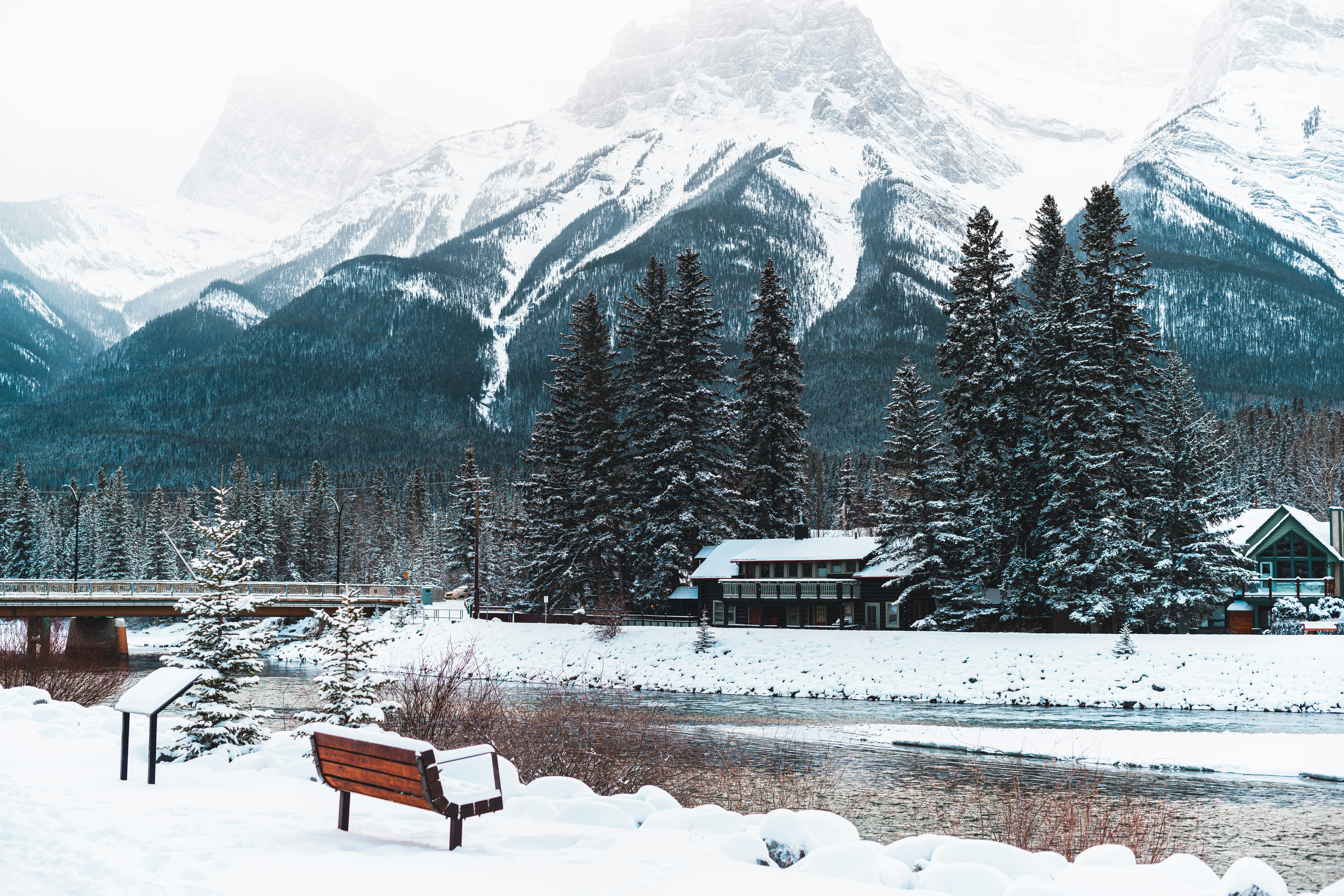 Park bench at the edge of a frozen river, with a snow-covered cabin and large mountain in the background