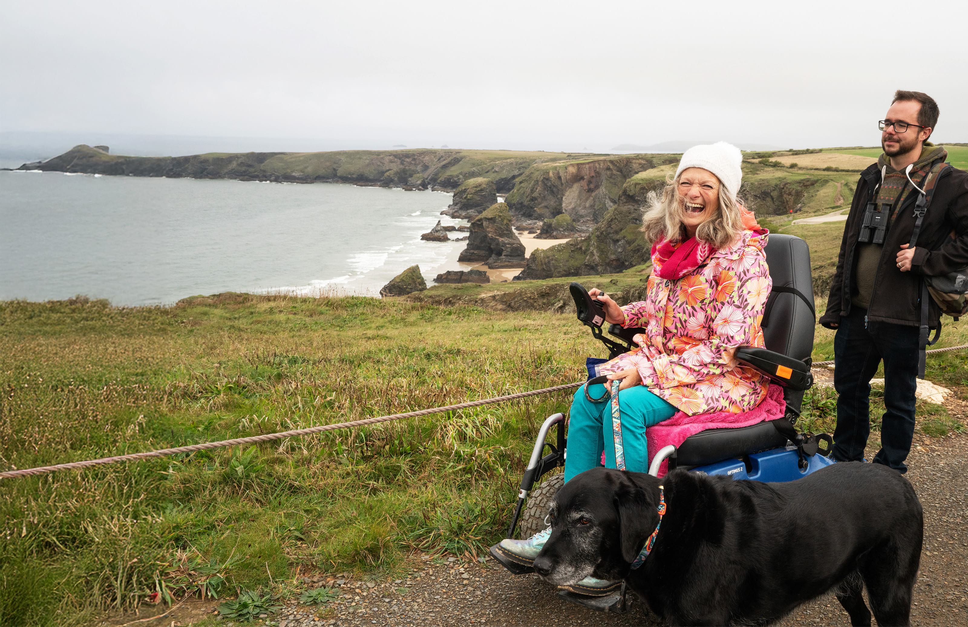 Lady in wheelchair and gentleman with their dog on coastal path looking out to sea
