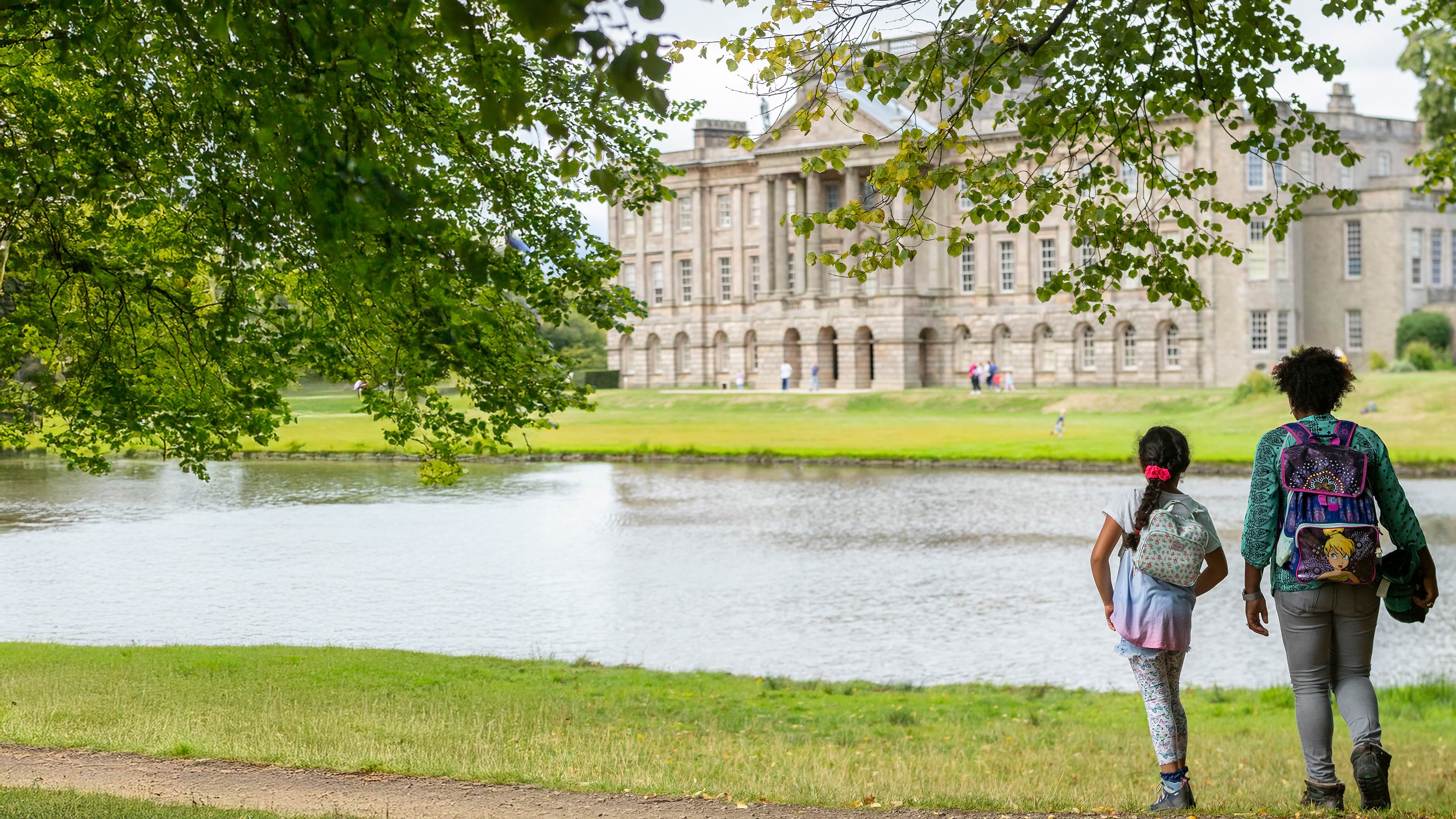 Family walking by water in parkland with view across to the very grand Lyme Park House