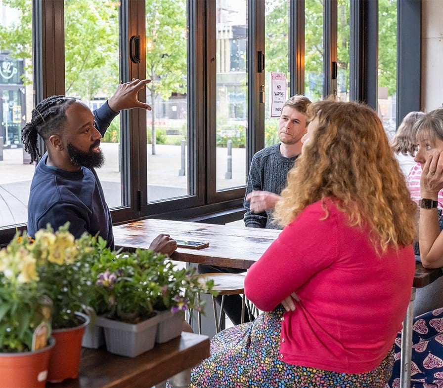 People participating in a workshop are listening to the facilitator whilst sat around a table with plants