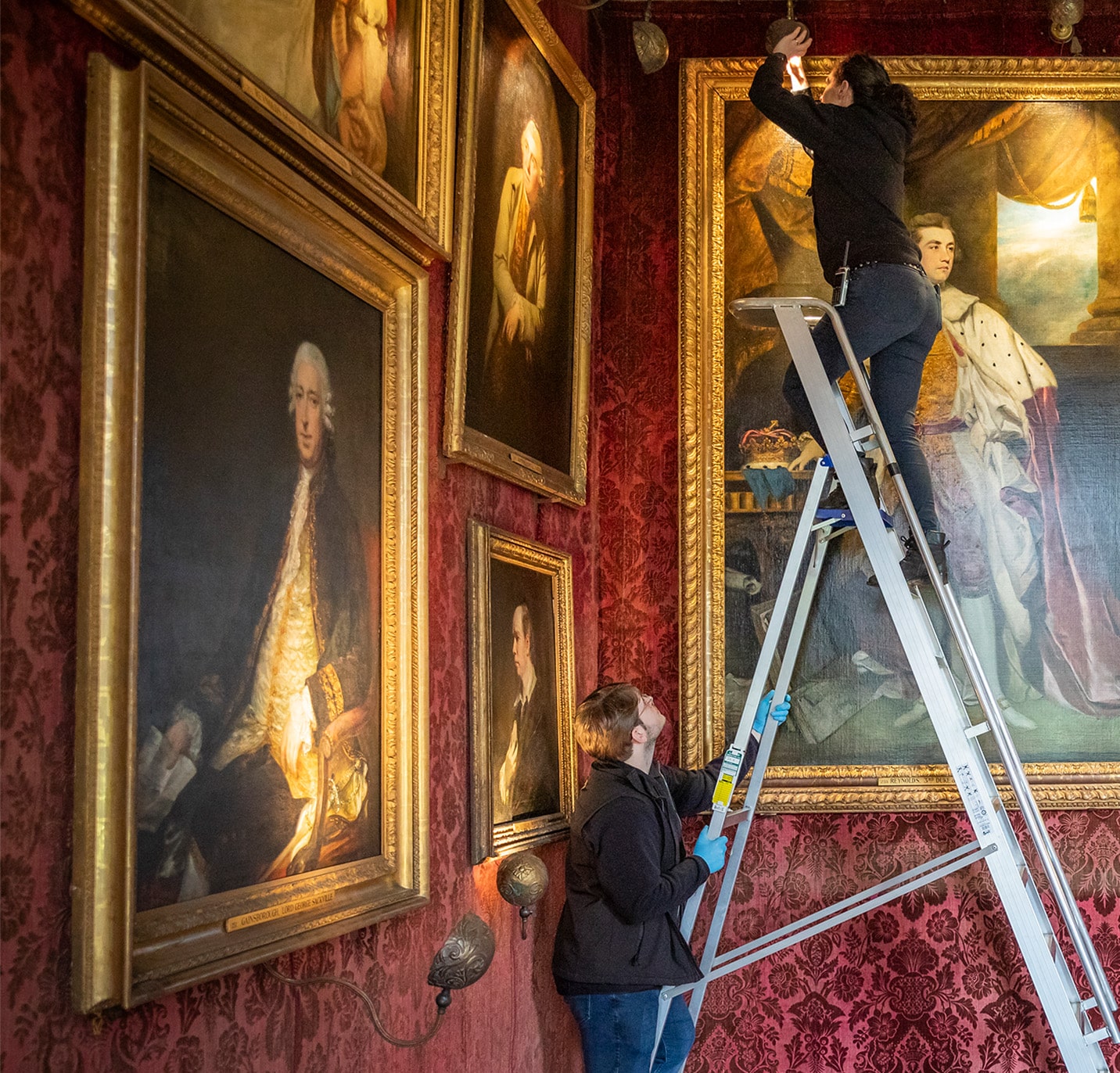 Conservators cleaning portraits and fitting lighting on a ladder