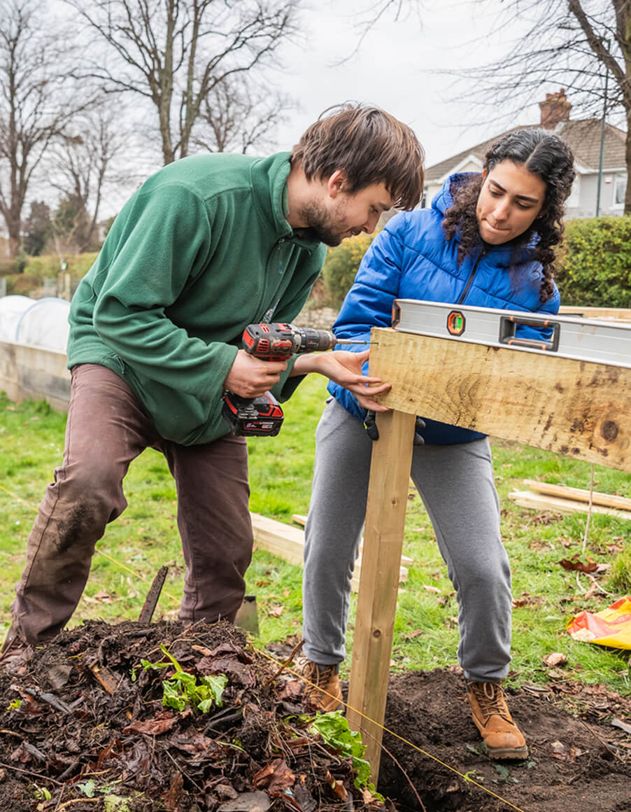 Two people building a compost bin. They are holding a piece of wood. There are raised beds in the background
