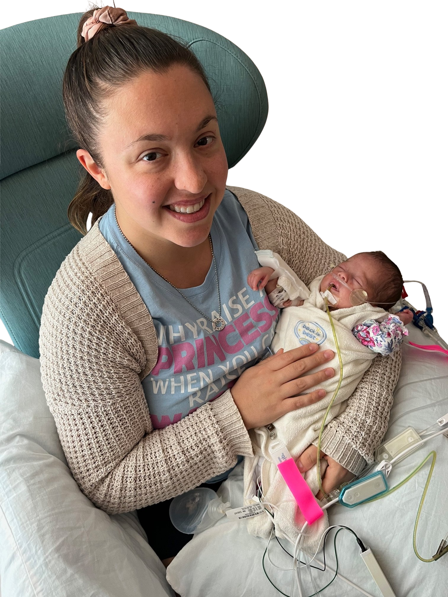 Person sitting in a chair, smiling at the camera while holding an infant wrapped in a blanket. The infant has medical tubes attached. The person is wearing a light blue shirt with partially visible text.
