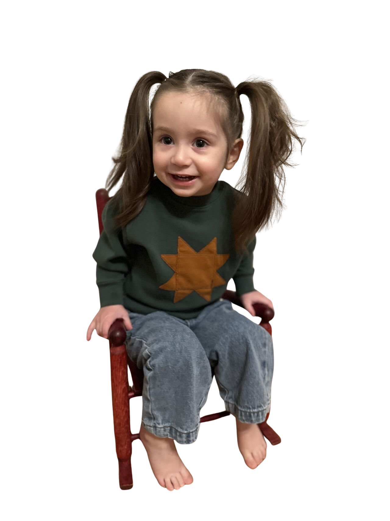 Young child sitting barefoot on a small red wooden chair, wearing a dark green sweater with an orange star design and light blue jeans. The child has long hair styled in two ponytails.