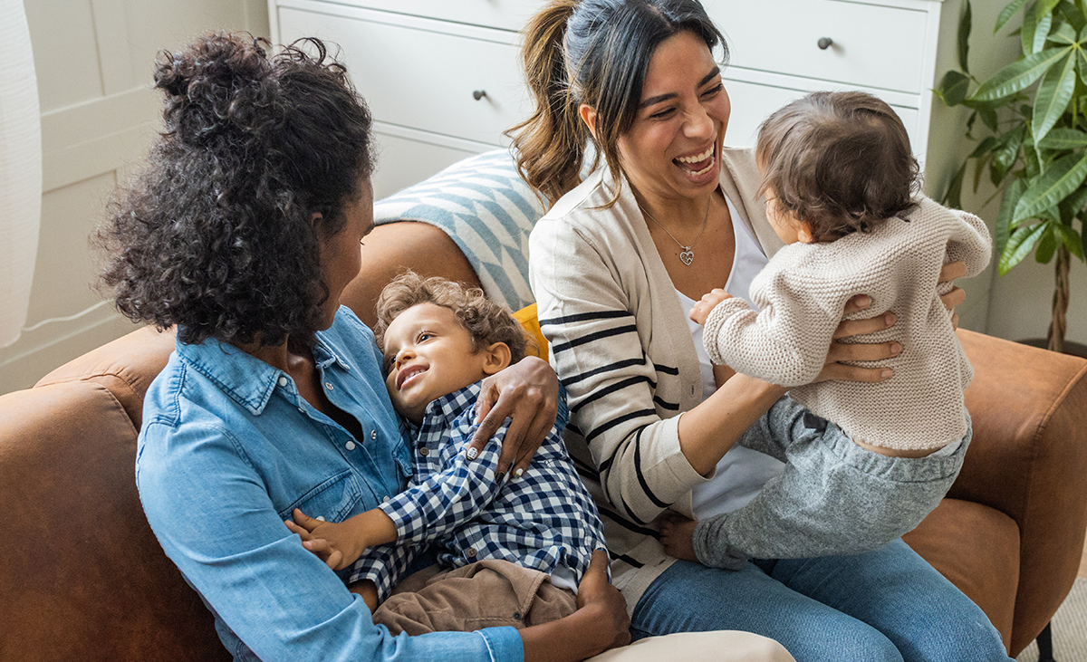 Two mothers on their couch laughing and playing with their children 