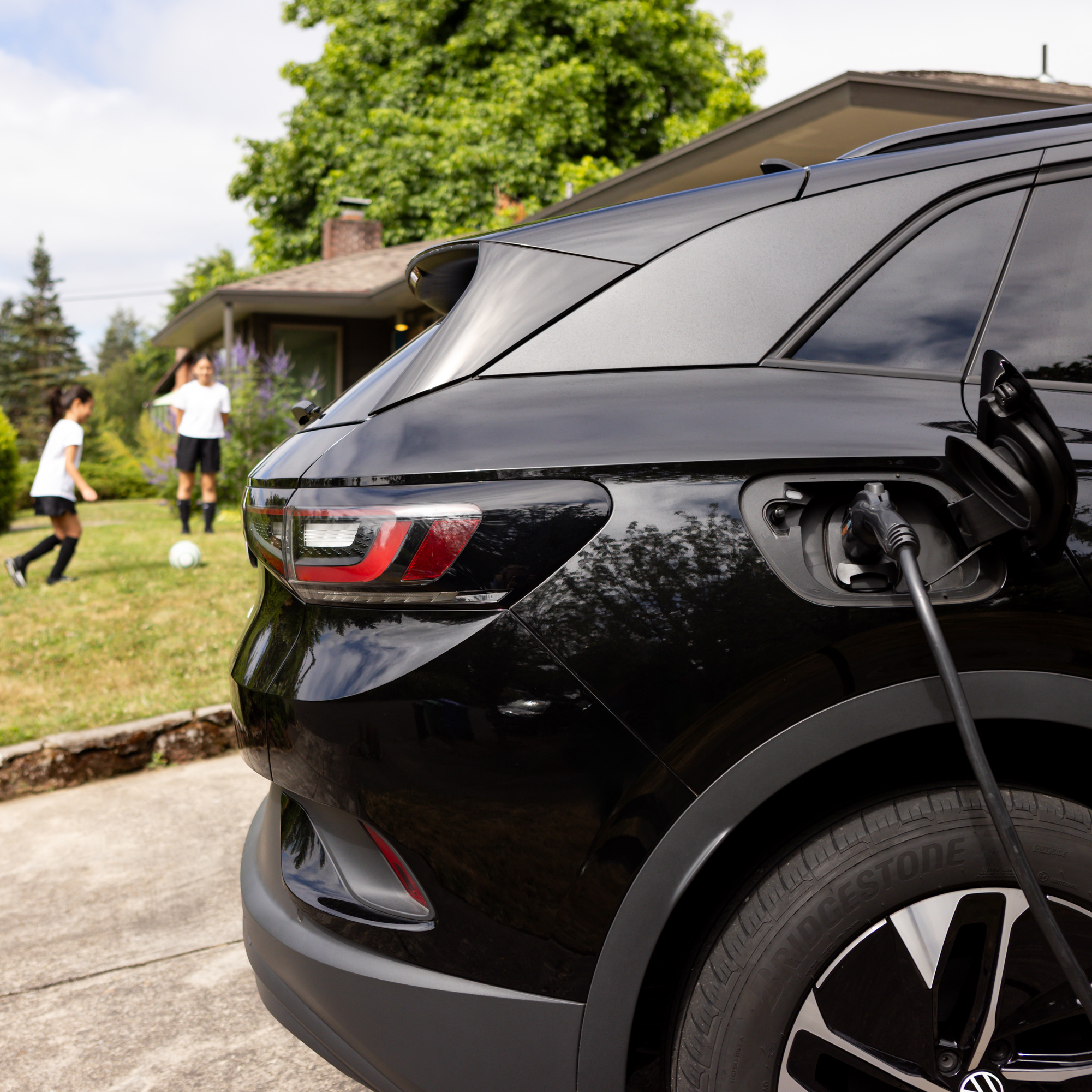 SUV charging in a driveway while two kids play soccer on a lush green lawn with blooming flowers. 