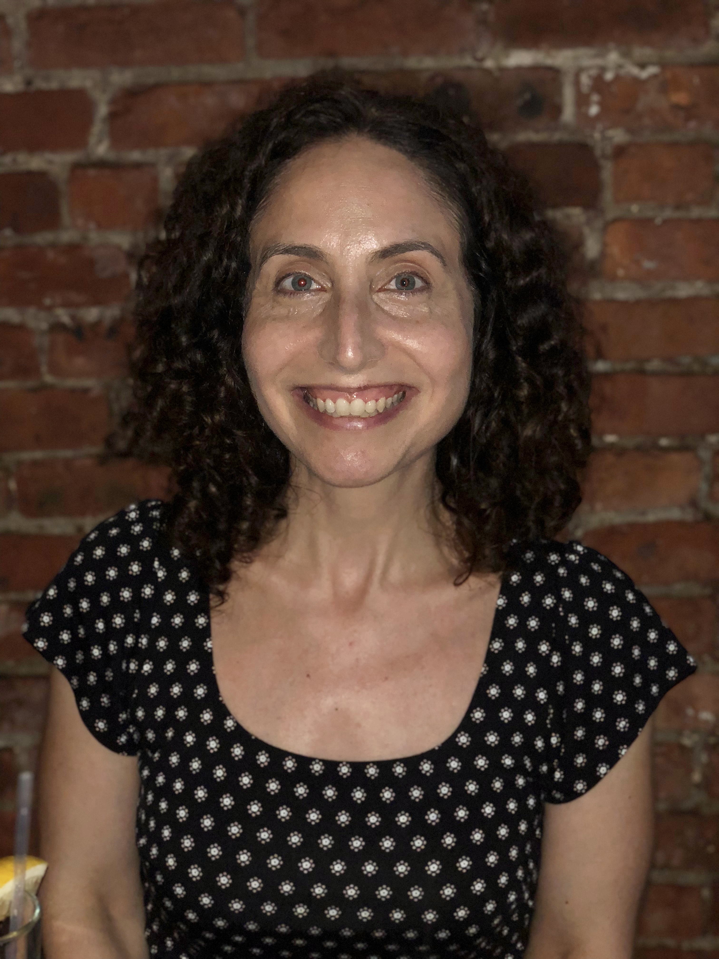The image shows a person with curly hair wearing a black dress with a white floral pattern, seated in front of a brick wall. A drink with a lemon wedge is visible on the table.