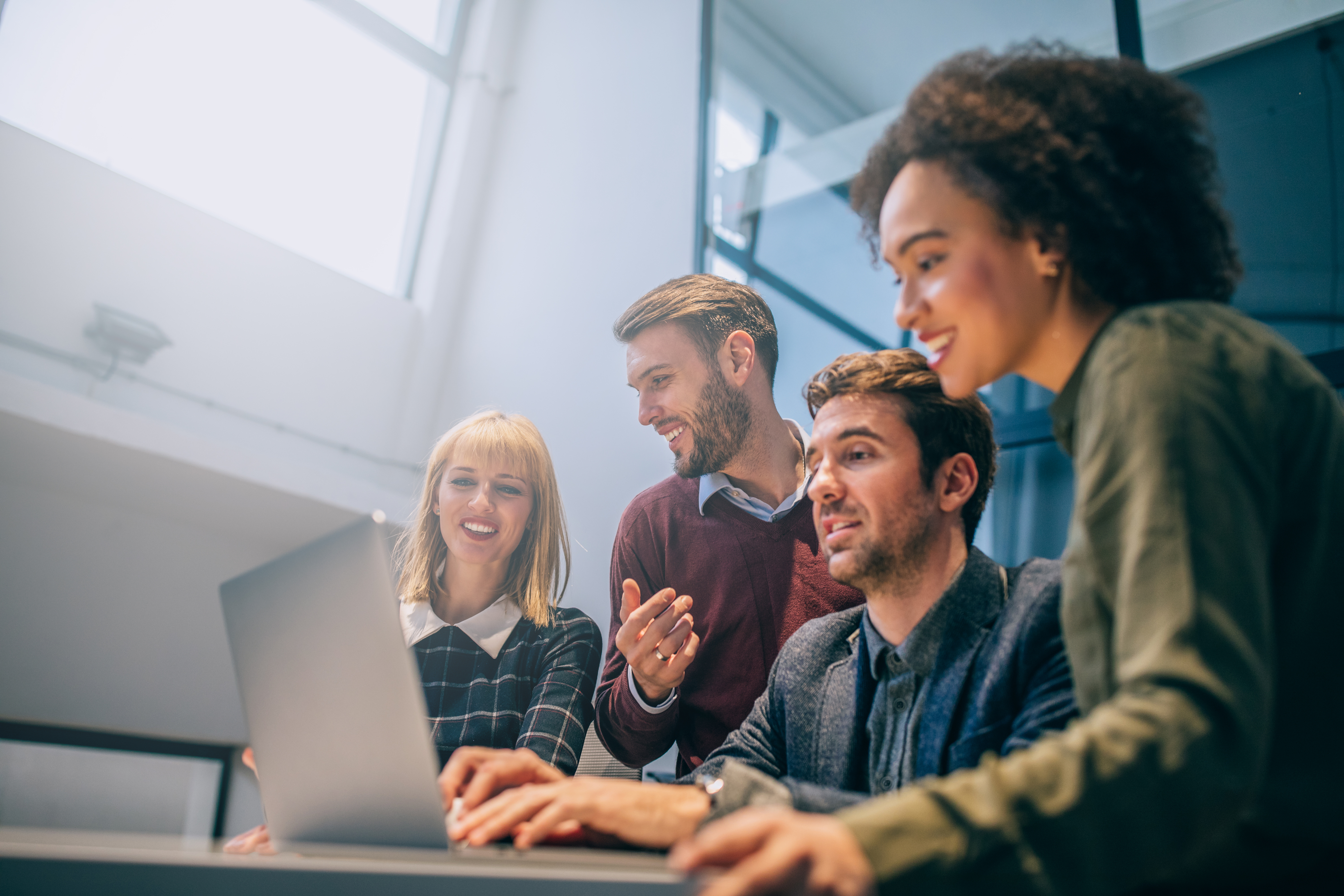 A group of four co-workers gathered around a laptop screen having a discussion and smiling 