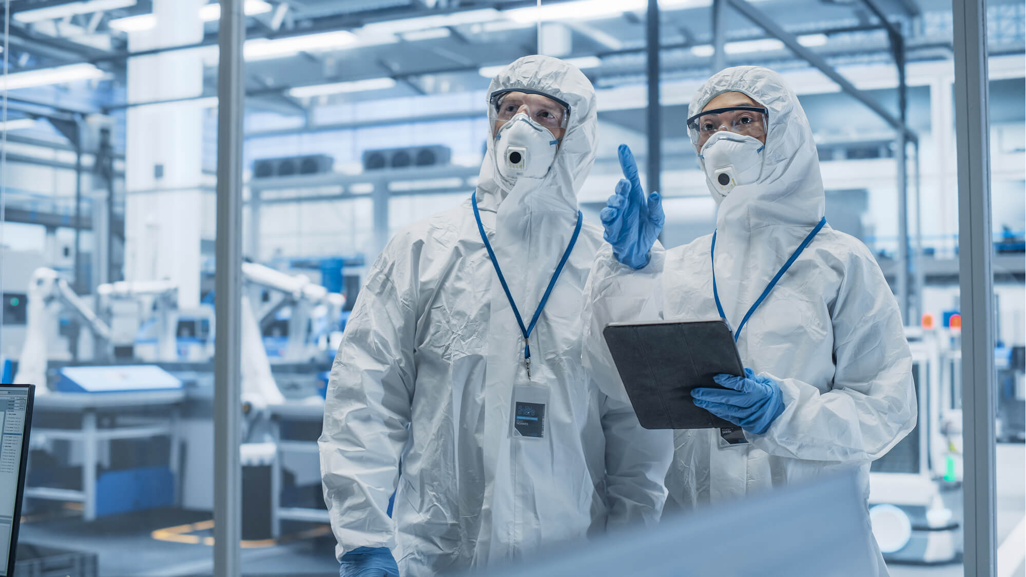 Two individuals wearing full protective suits and gloves, possibly in a cleanroom or laboratory setting. They appear to be discussing or reviewing information on a tablet device, indicating a focus on precision and safety in a controlled environment. The background suggests a high-tech or industrial facility, with machinery and equipment visible.