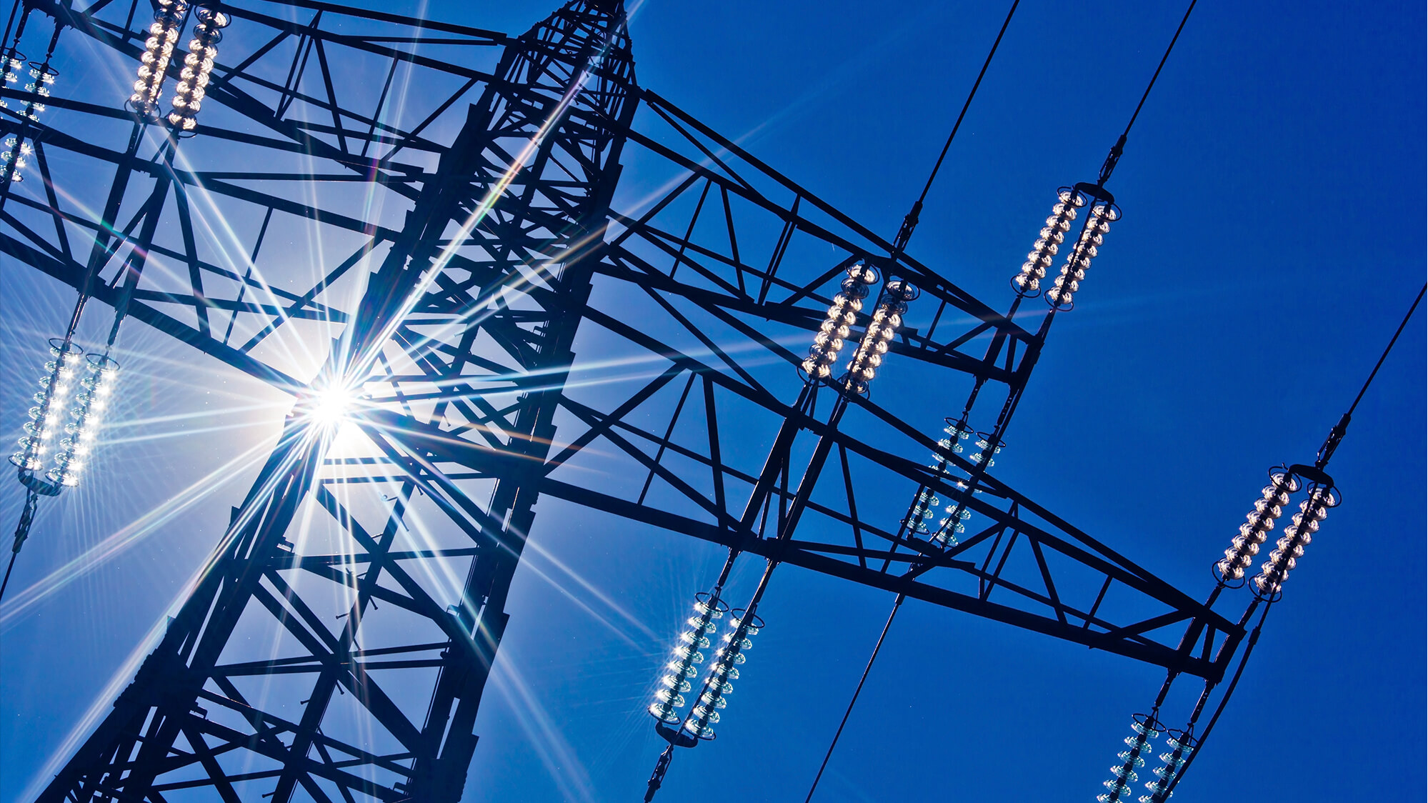 A high-voltage transmission tower, commonly used in power grids to transport electricity over long distances. The sun is shining brightly in the background, highlighting the structure's intricate network of wires and insulators. This setup is essential for efficiently distributing electrical power from generation sources to consumers, ensuring a reliable energy supply.