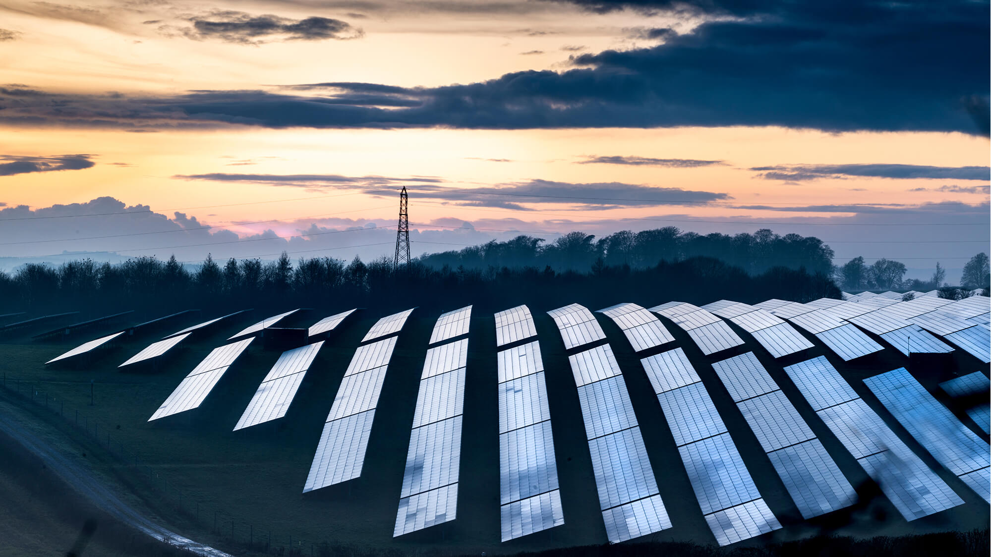 A solar farm at sunset, showcasing rows of solar panels on a field. The panels are positioned to capture sunlight efficiently, and the scene is set against a backdrop of trees and a cloudy sky, with an electricity pylon visible in the distance. This setting highlights the integration of renewable energy technology within a natural landscape.