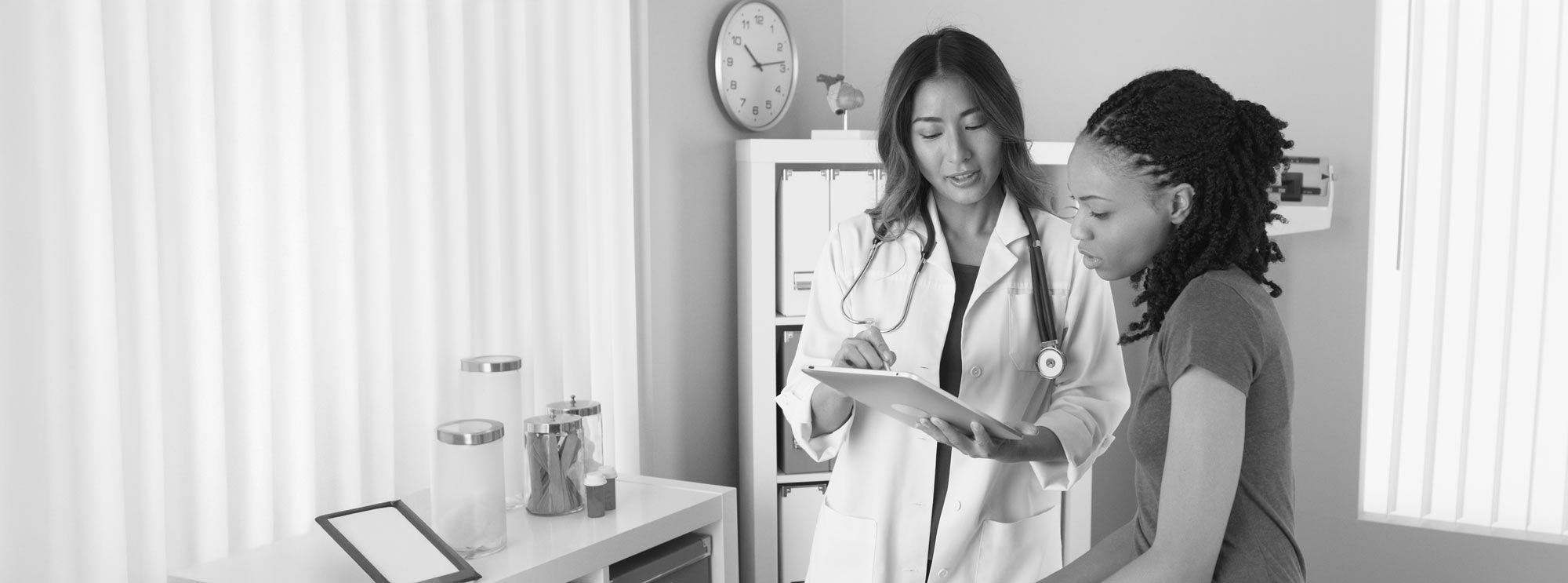 The image depicts a medical consultation between a healthcare professional and a patient. The healthcare professional is wearing a white coat and has a stethoscope, indicating a medical setting. They are using a tablet, possibly for reviewing medical records or taking notes. The environment is a clinical office with medical supplies visible, such as jars and a scale.