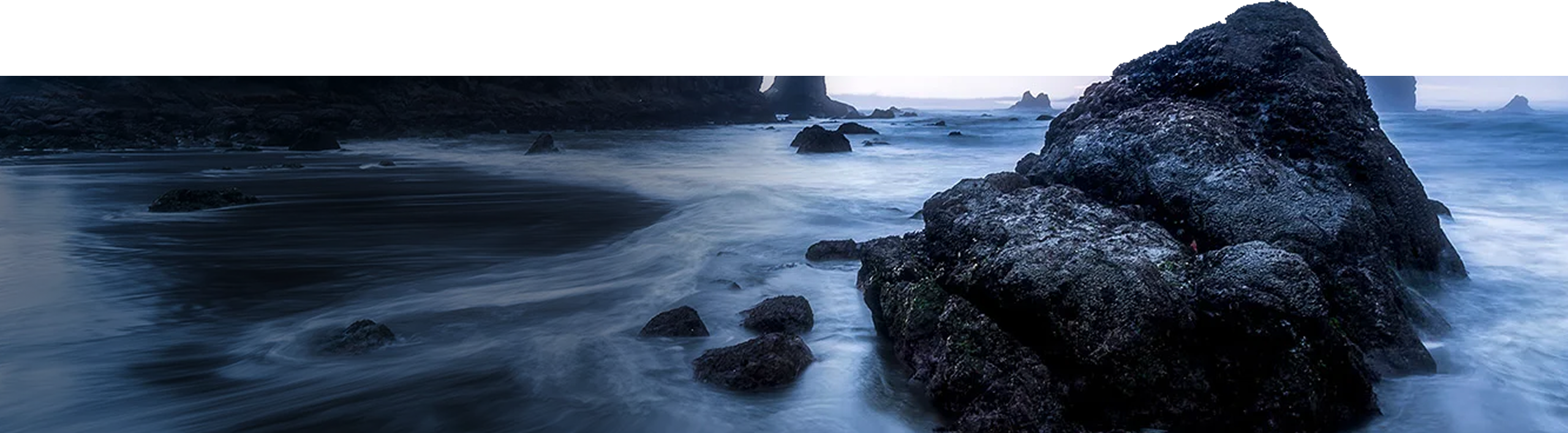 Rocky shoreline with large boulders and misty waves under a dim, blue-toned sky.
