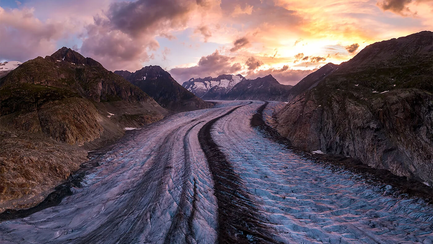 A winding glacier surrounded by rugged mountains at sunset with colorful clouds overhead.