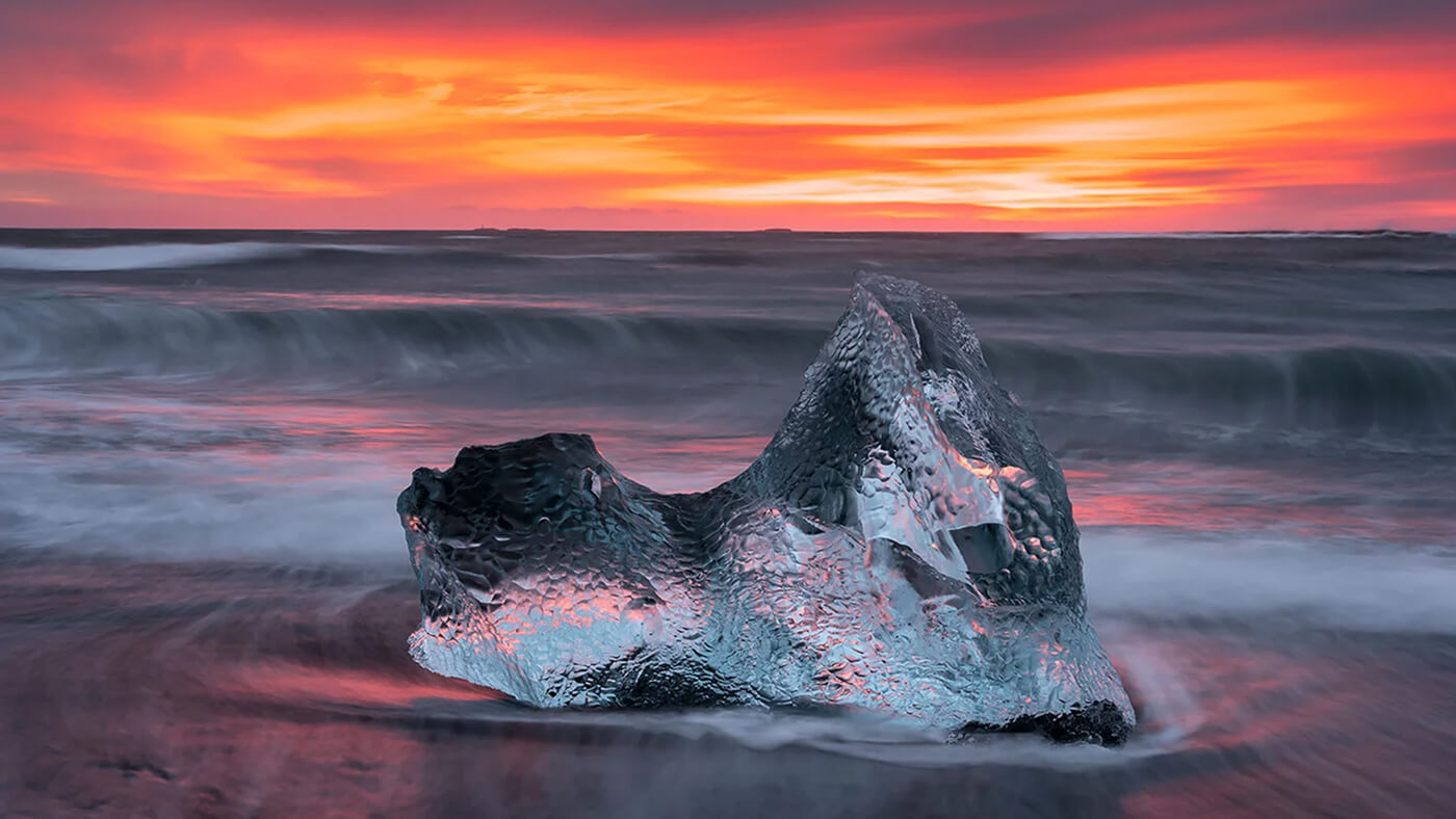 A large, clear ice chunk on a beach with waves and a vibrant orange sunset sky in the background.