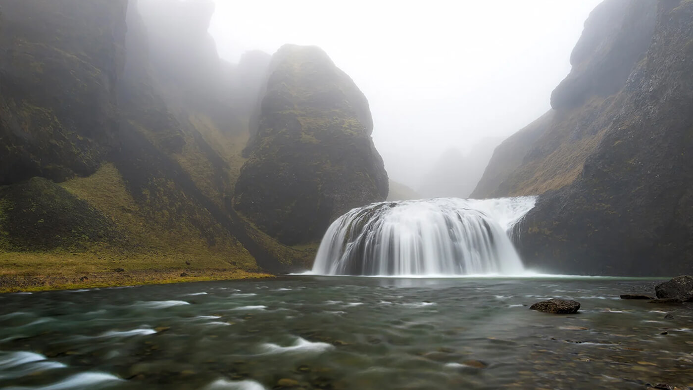 A wide waterfall cascading between mossy cliffs in a misty, mountainous landscape.