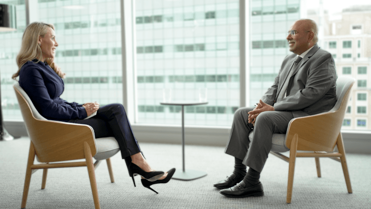 Two people seated in armchairs having a filmed conversation, with a city office skyline visible through large windows behind them.