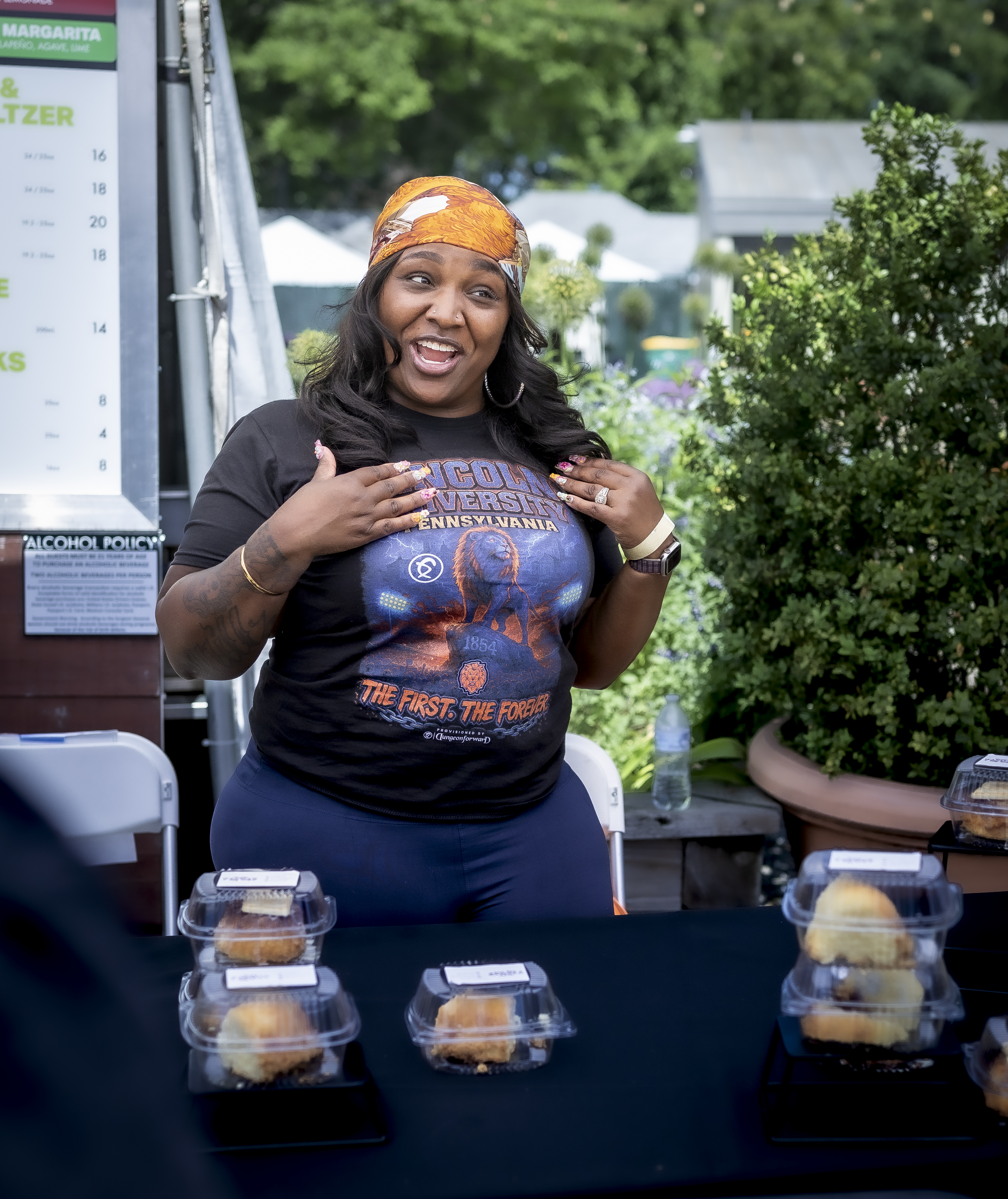Tasha (an African American woman with black hair, wearing a black t-shirt and jeans), smiling and laughing in front of a table of her goods she's selling
