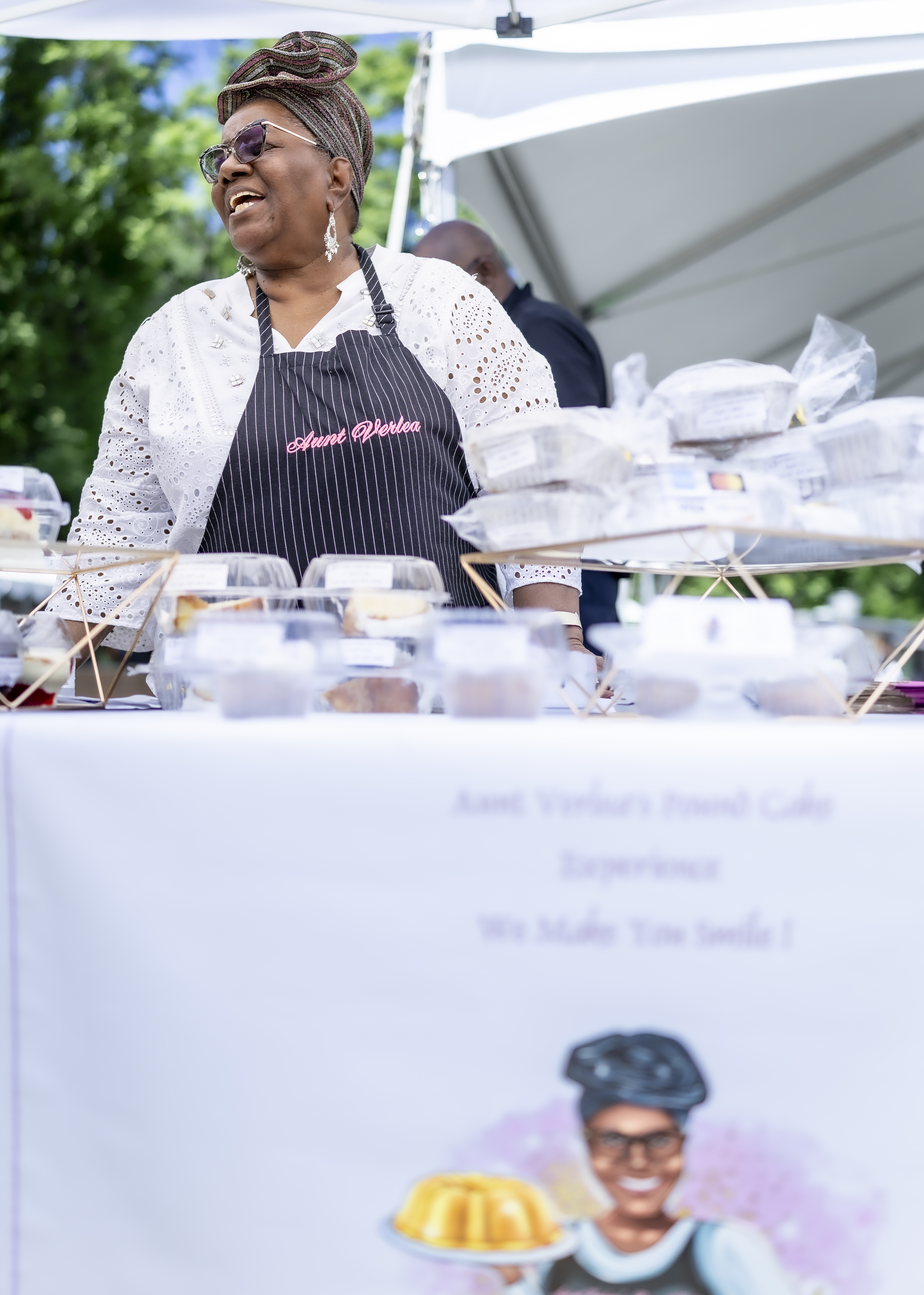 Romona (an African American woman wearing sunglasses and a black apron) laughing in front of her pastries