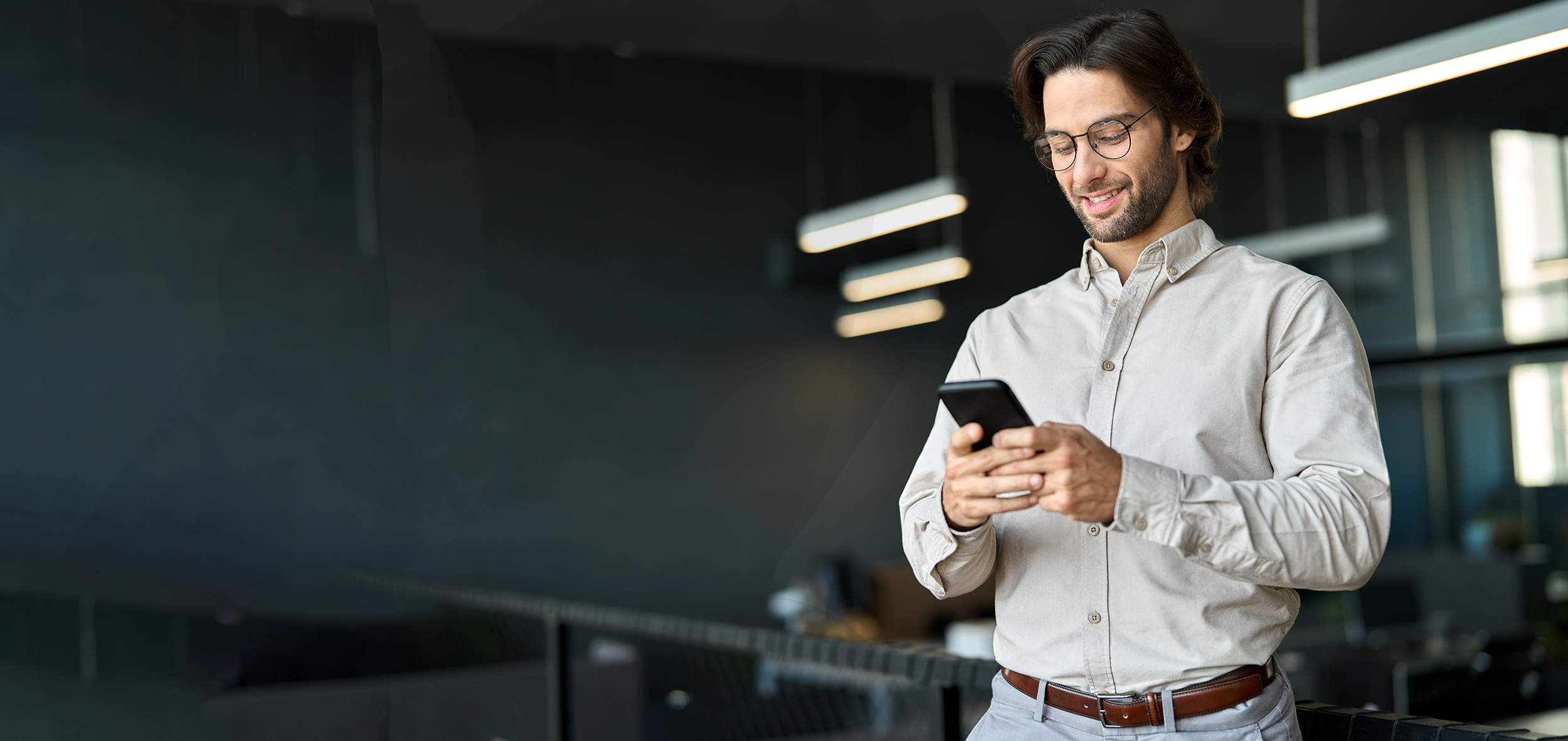 Man with brown hair, beard, and glasses looking at phone at work 