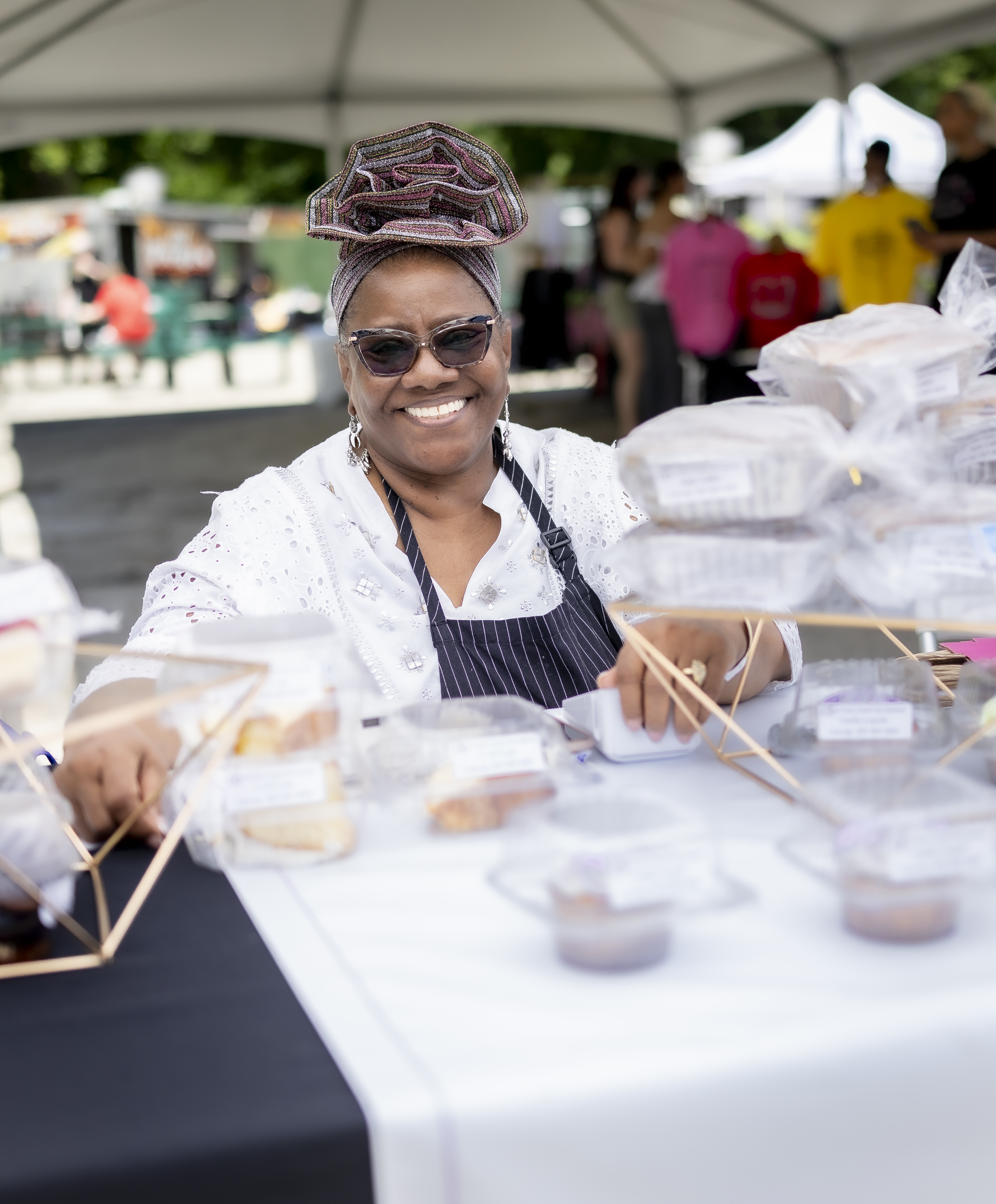Romona (an African American woman wearing sunglasses and a black apron) laughing in front of her pastries