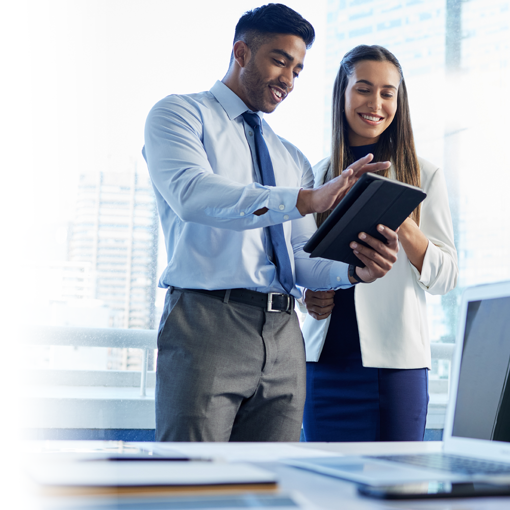 A smiling young man and woman stand in an office looking at an iPad together.