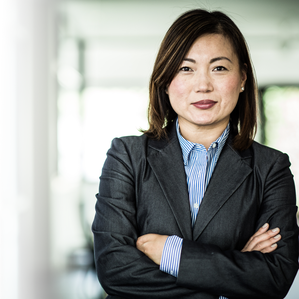 An professional Asian woman stands in an office. Her arms are crossed and her face has an expression of confidence and determination. 