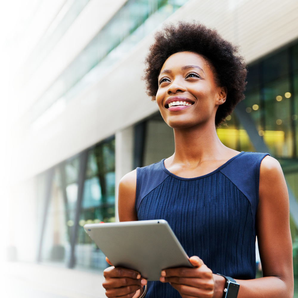 A young black woman stands outdoors in front of an office holding an iPad. Her face is uplifted and she is smiling.
