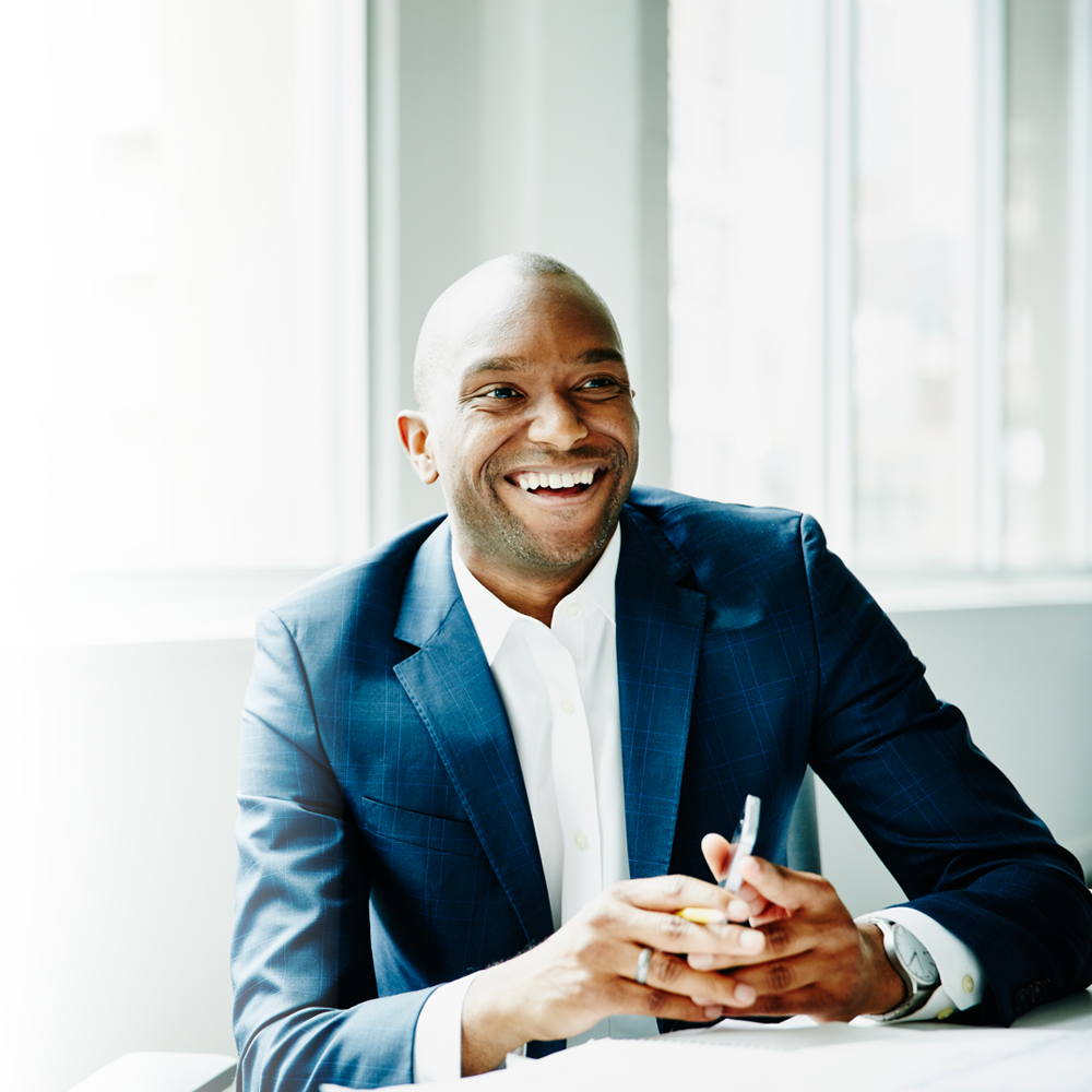 A smiling black man sits in an office holding a phone. He's wearing a blue suit. 