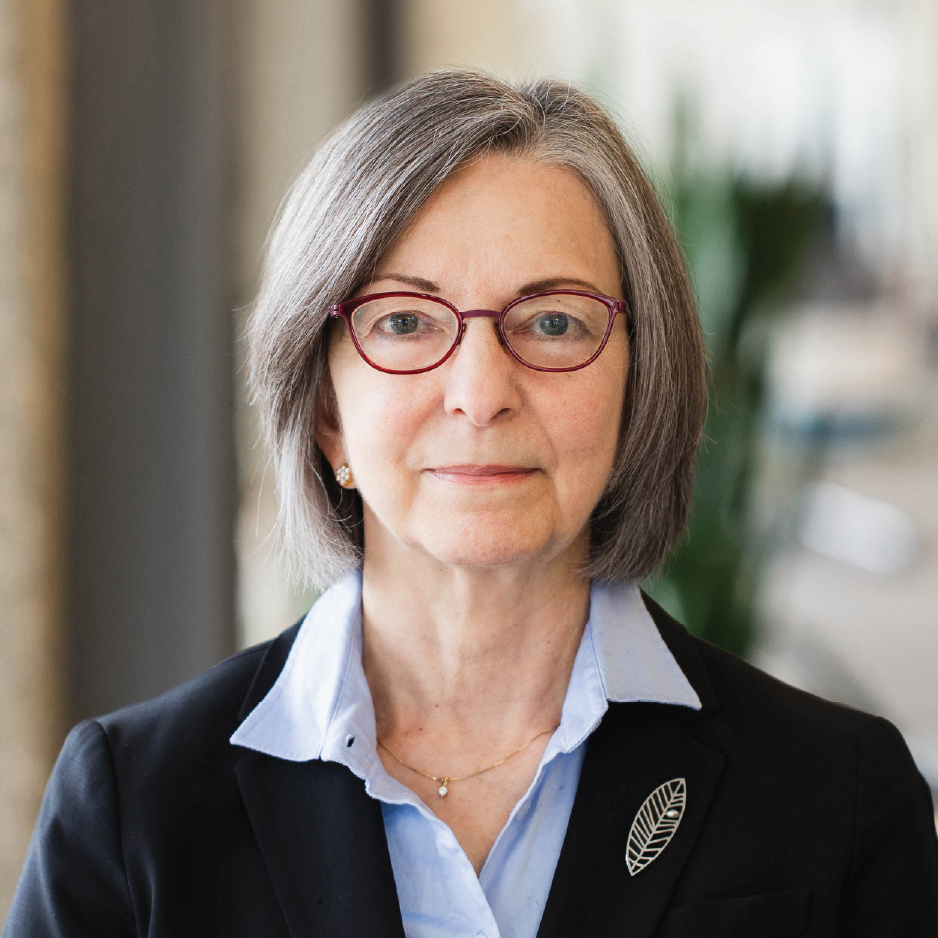 Beth Namachchivaya wearing a black blazer with blue collared shirt, brown glasses with short grey-brown hair in an indoor setting with blurred background