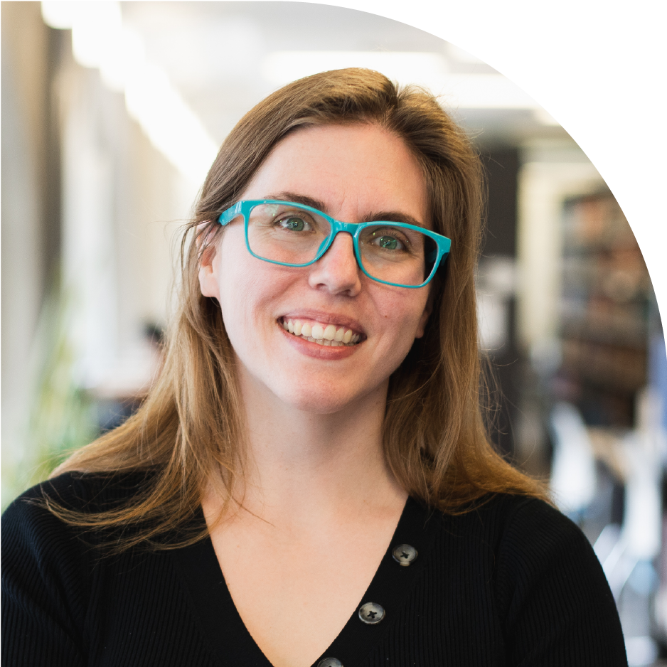 Lauren Byl wearing a black shirt, blue-green glasses with long brown hair in an indoor setting with blurred background