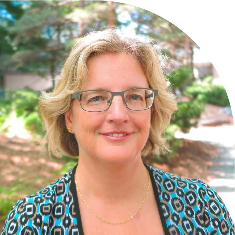 Jackie Stapleton with short blonde hair standing outdoors in a garden setting wearing a blue patterned blouse and grey glasses