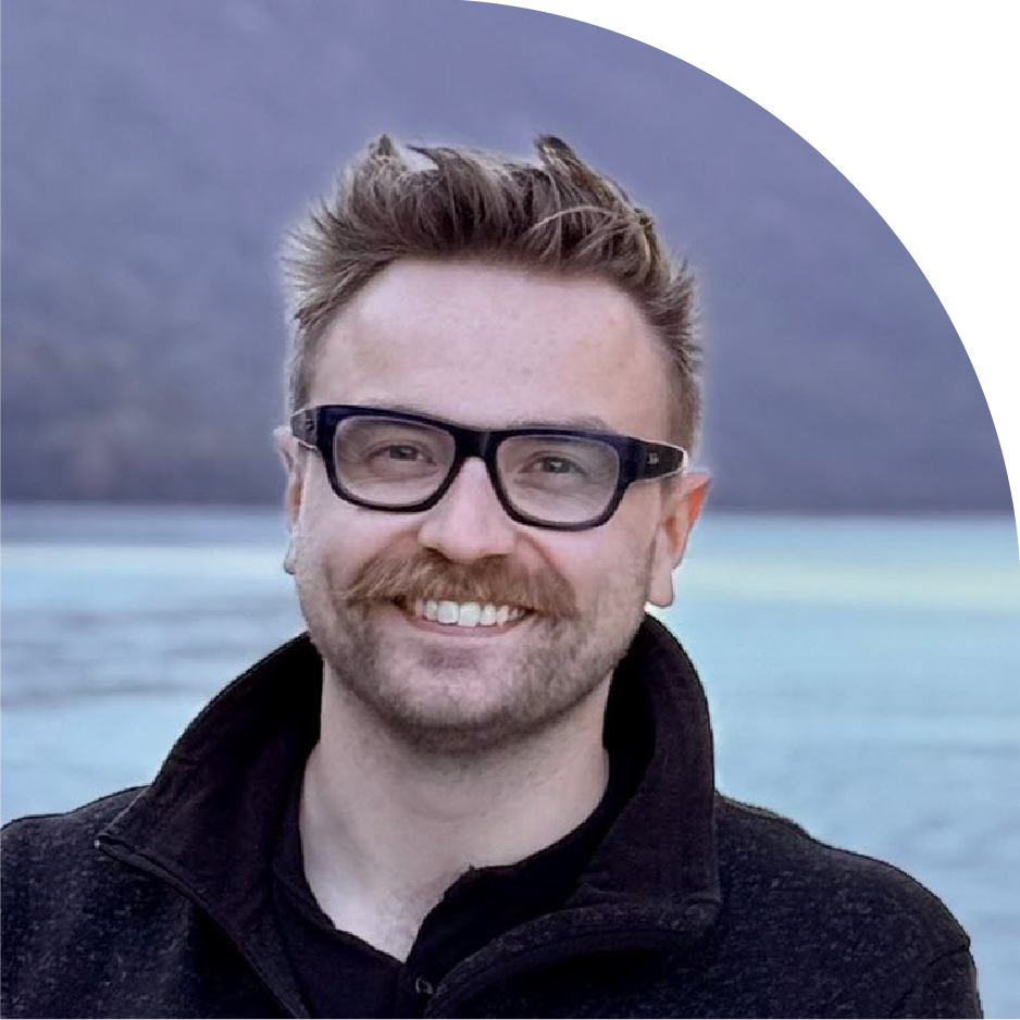 Ryan Ball with short brown hair, black glasses wearing a black jacket in an outdoor setting with a body of water in the background